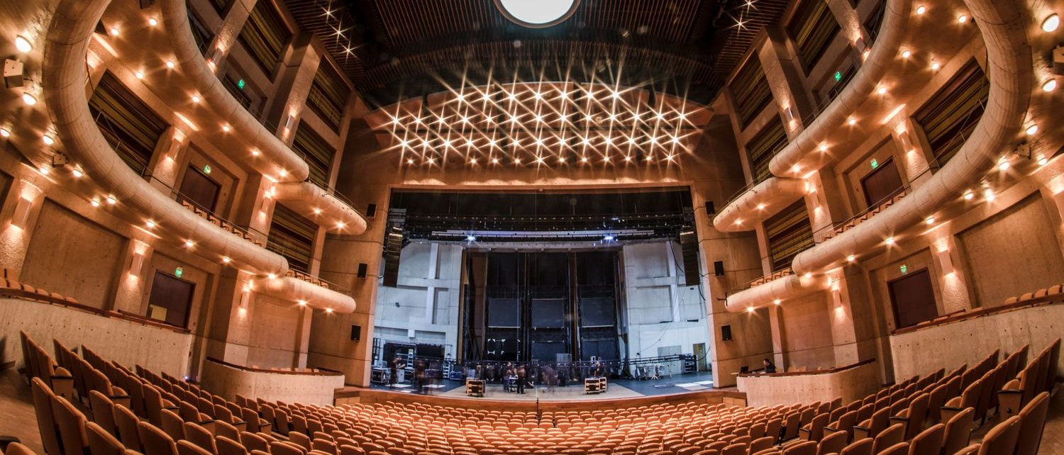A wide-angle view of an empty modern theater auditorium with rows of tan seats, curved balconies, and a large stage under bright ceiling lights.