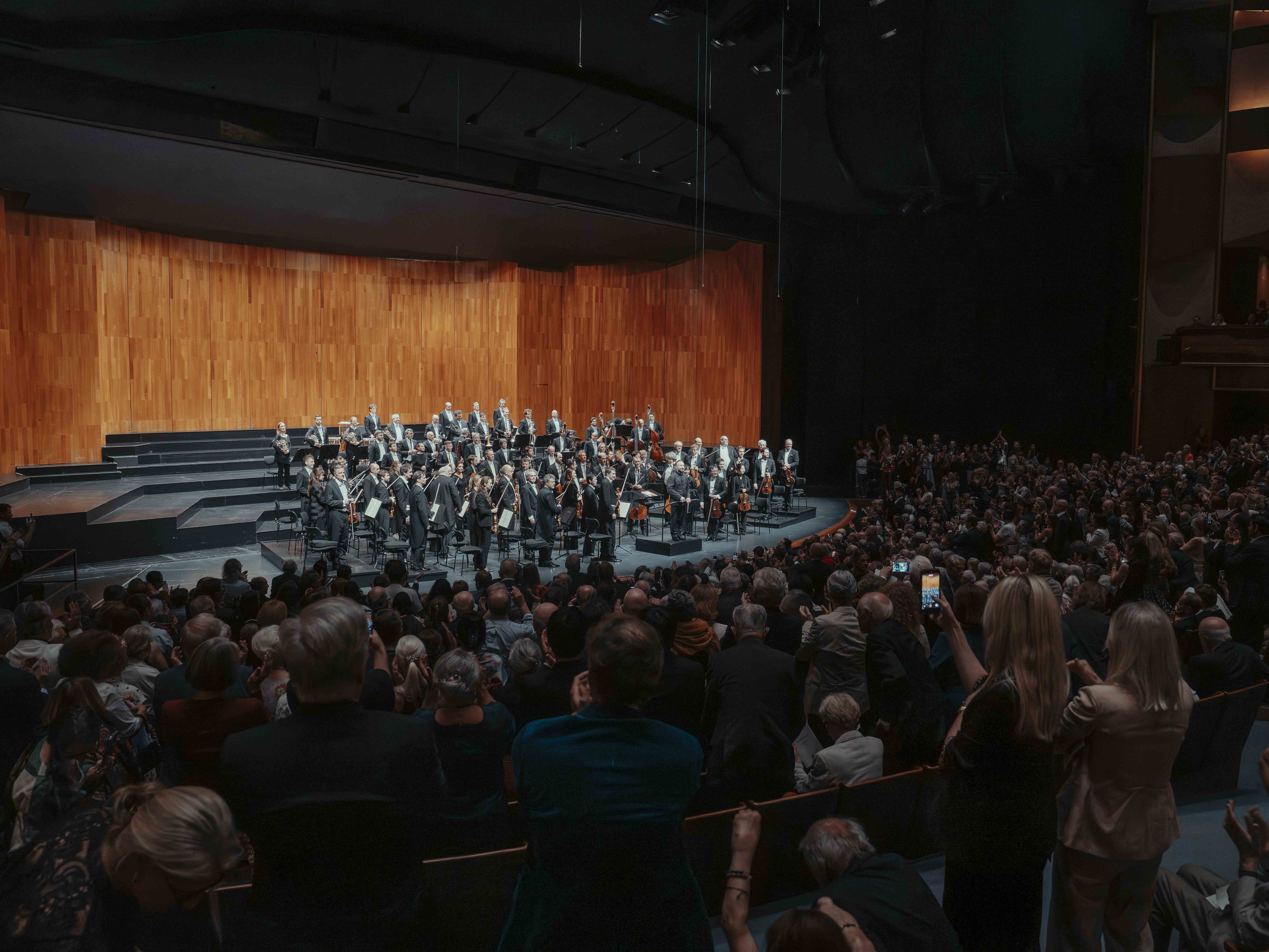 Ein Sinfonieorchester steht auf der Bühne und blickt auf ein großes, applaudierendes Publikum in einem Konzertsaal mit Holzvertäfelung und gedämpfter Beleuchtung. Einige Zuschauer machen Fotos oder Videos.