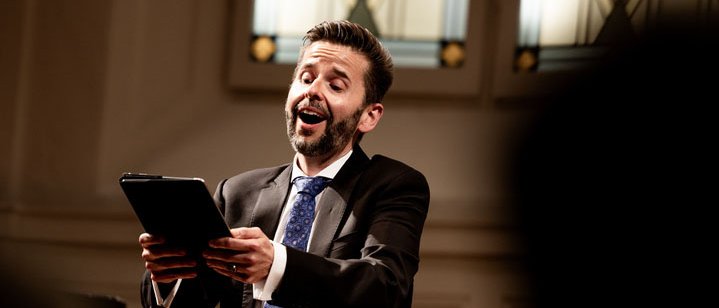 A man in a suit sings passionately while holding a tablet in a well-lit room with large stained glass windows behind him. An audience is visible in the foreground.