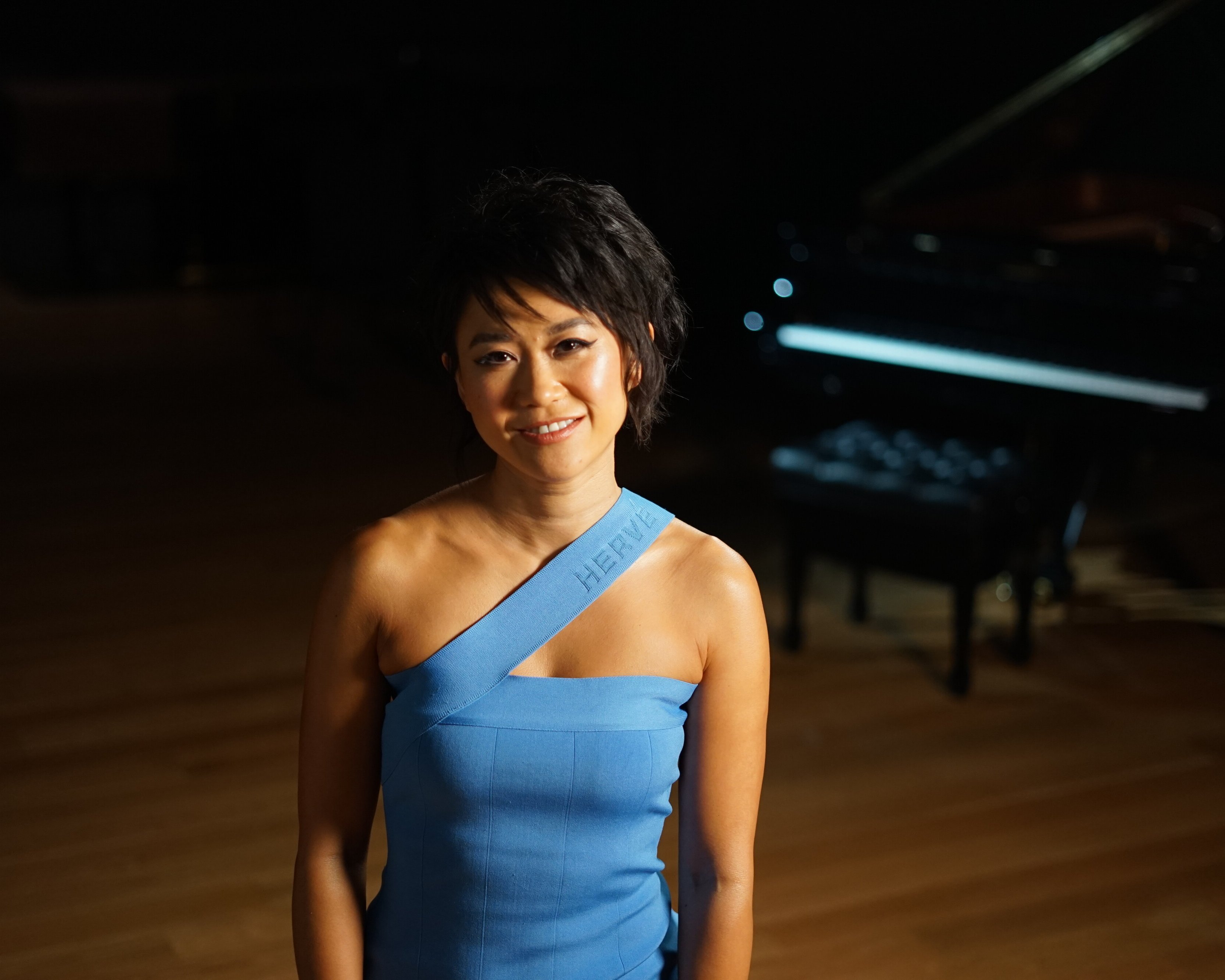 Yuja Wang in einem blauen, schulterfreien Kleid steht lächelnd in einem sanft beleuchteten Raum mit einem Flügel im Hintergrund.