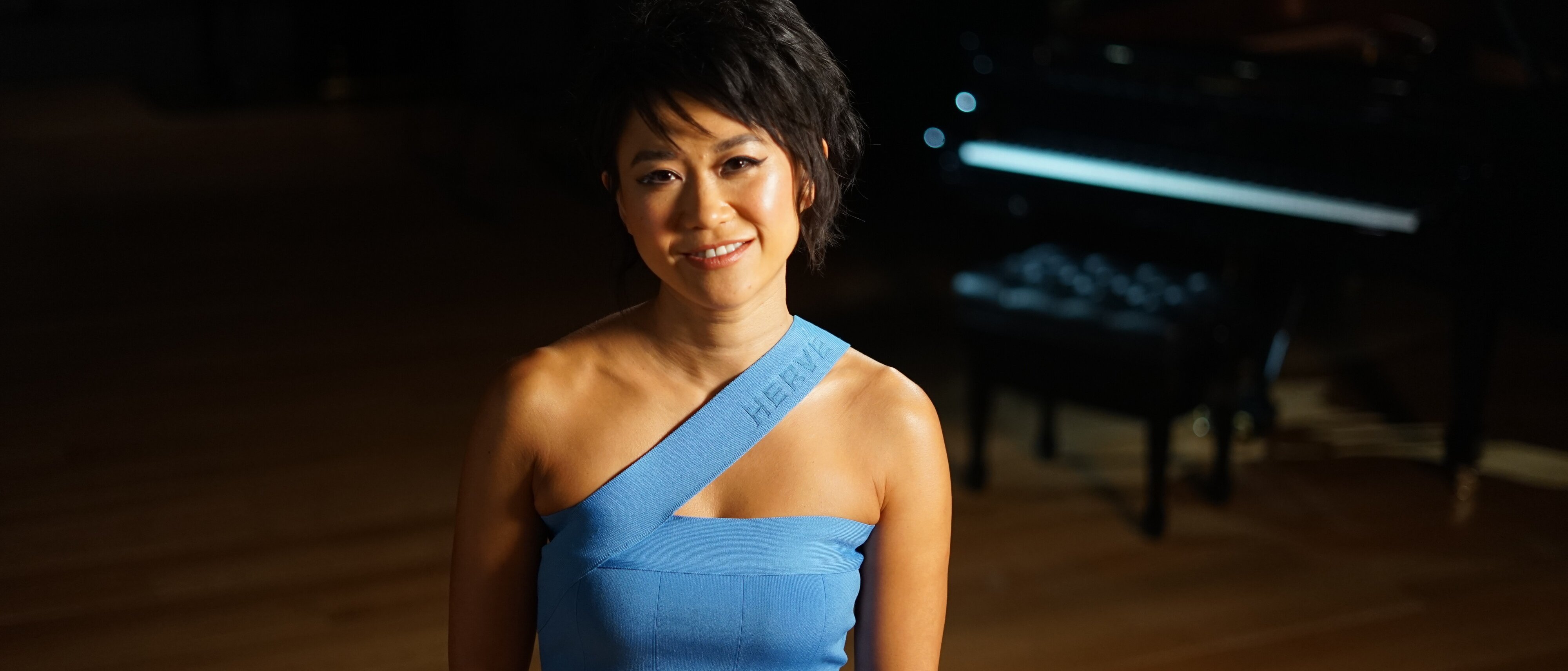 Yuja Wang in einem blauen, schulterfreien Kleid steht lächelnd in einem sanft beleuchteten Raum mit einem Flügel im Hintergrund.
