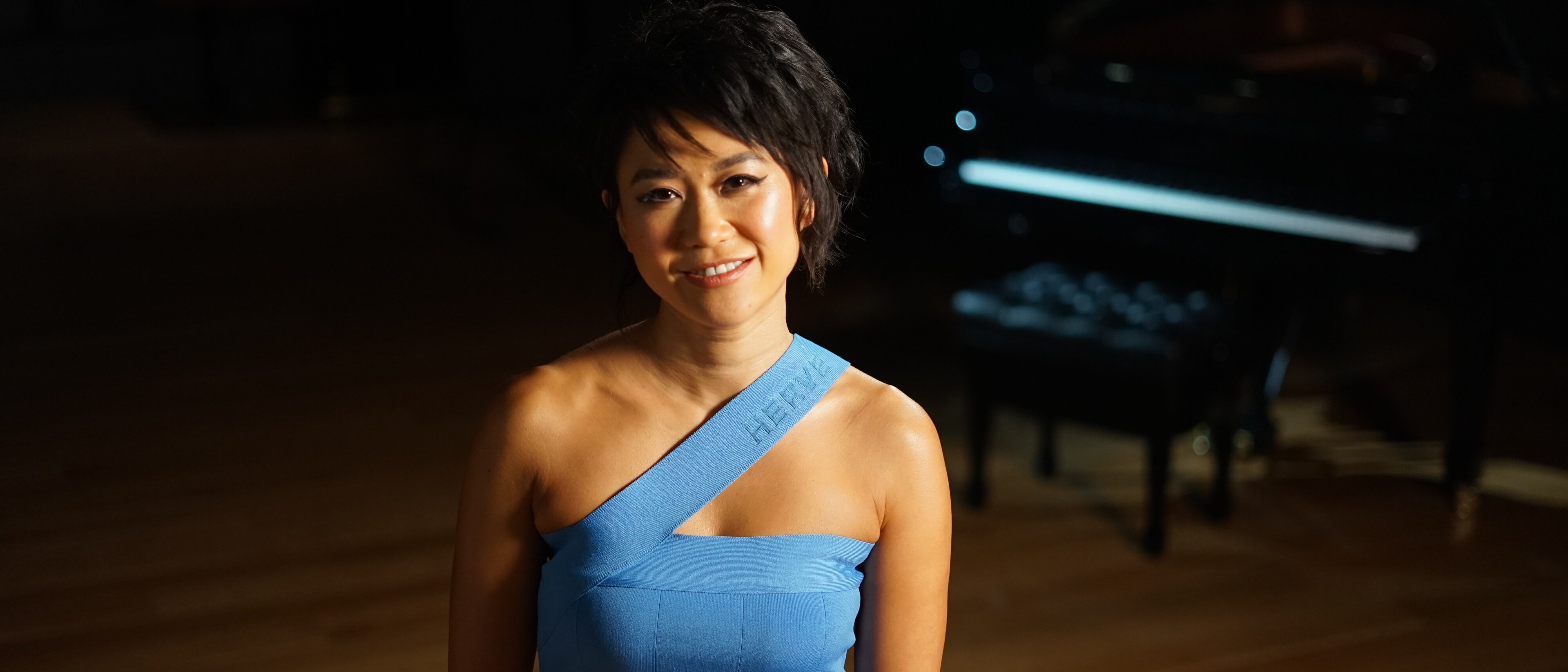Yuja Wang in einem blauen, schulterfreien Kleid steht lächelnd in einem sanft beleuchteten Raum mit einem Flügel im Hintergrund.