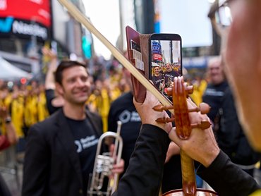 Ein Musiker hält ein Smartphone in der Hand, um ein Foto von zwei anderen Musikern am Times Square zu machen, während er in der anderen Hand einen Geigenbogen hält.