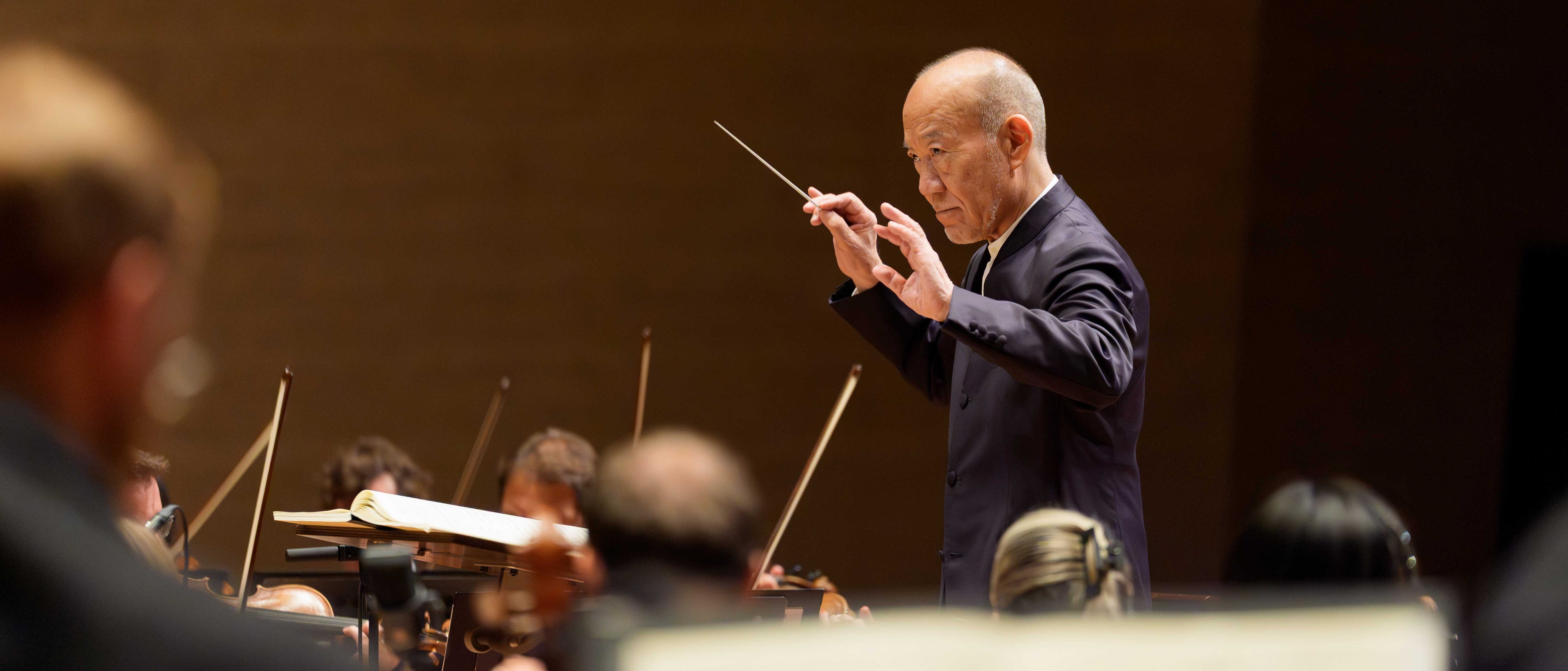 Ein Dirigent hält einen Taktstock und leitet ein Orchester während einer Aufführung. Im Vorder- und Hintergrund sind Musiker mit Streichinstrumenten zu sehen. Der Schauplatz scheint ein Konzertsaal zu sein.