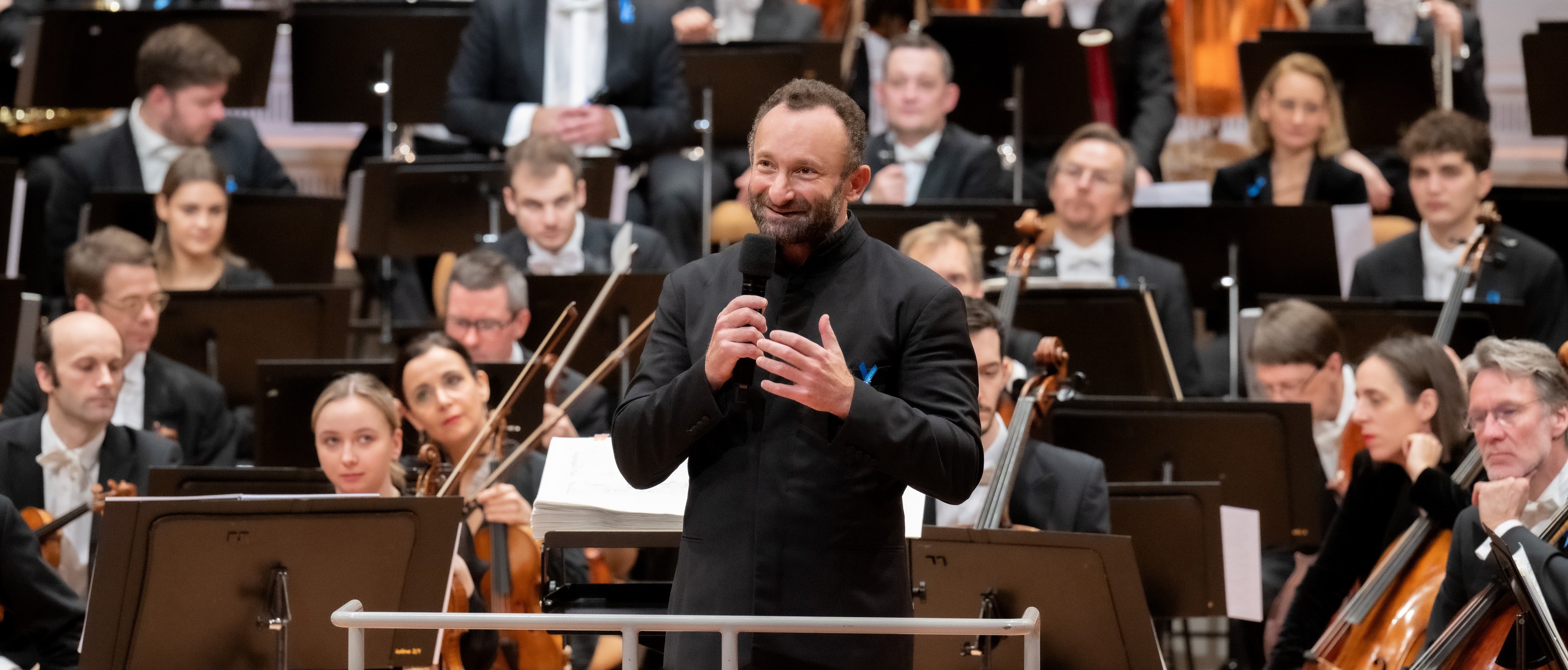 Ein Dirigent steht an einem Podium und spricht in ein Mikrofon, während hinter ihm auf der Bühne ein ganzes Orchester sitzt. Die Musiker halten Instrumente, und im Hintergrund ist ein geschmückter Baum zu sehen.