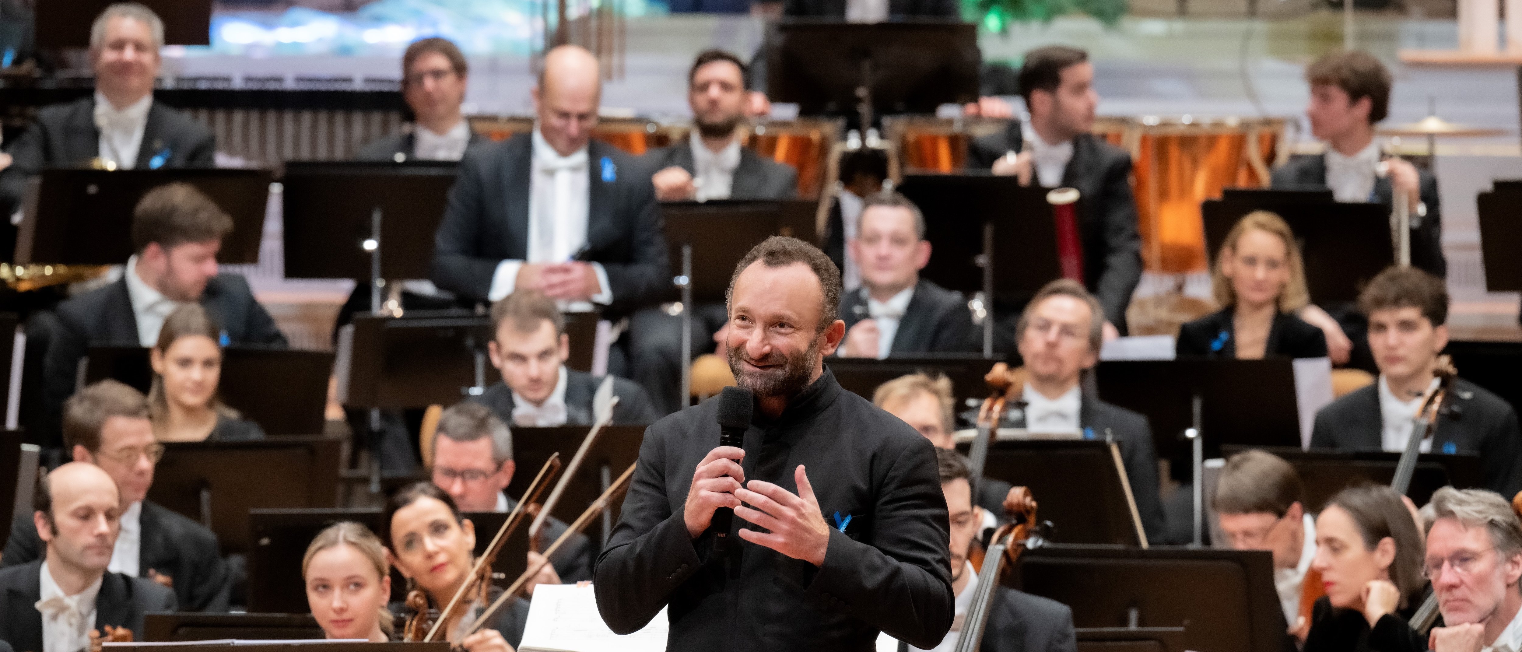 A conductor speaks into a microphone on stage in front of an orchestra, whose musicians sit with their instruments and music stands during a formal concert performance.
