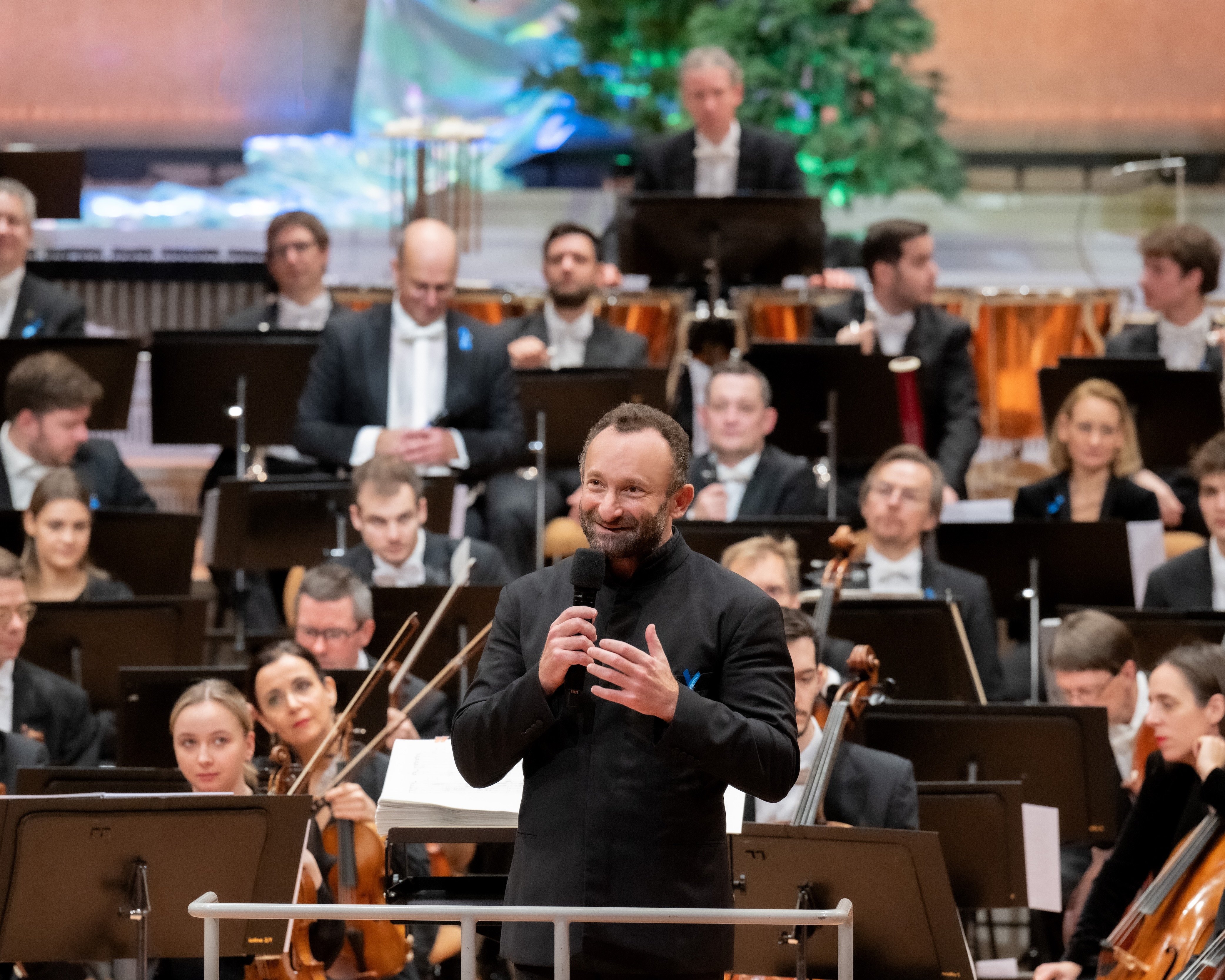 Ein Dirigent steht an einem Podium und spricht in ein Mikrofon, während hinter ihm auf der Bühne ein ganzes Orchester sitzt. Die Musiker halten Instrumente, und im Hintergrund ist ein geschmückter Baum zu sehen.