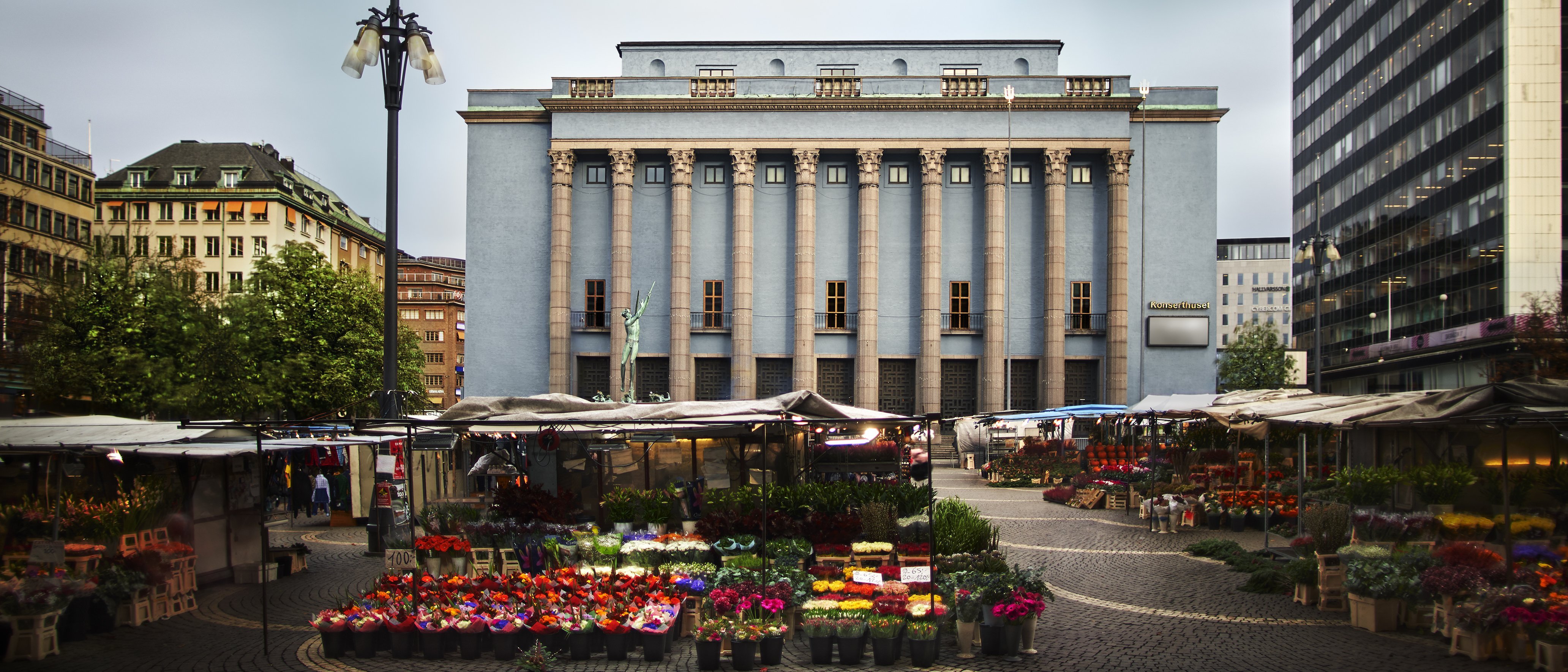 Ein Blumenmarkt im Freien mit bunten Ständen auf einem Stadtplatz, umgeben von modernen und historischen Gebäuden, mit einem großen, blauen neoklassizistischen Gebäude in der Mitte im Hintergrund unter einem bewölkten Himmel.