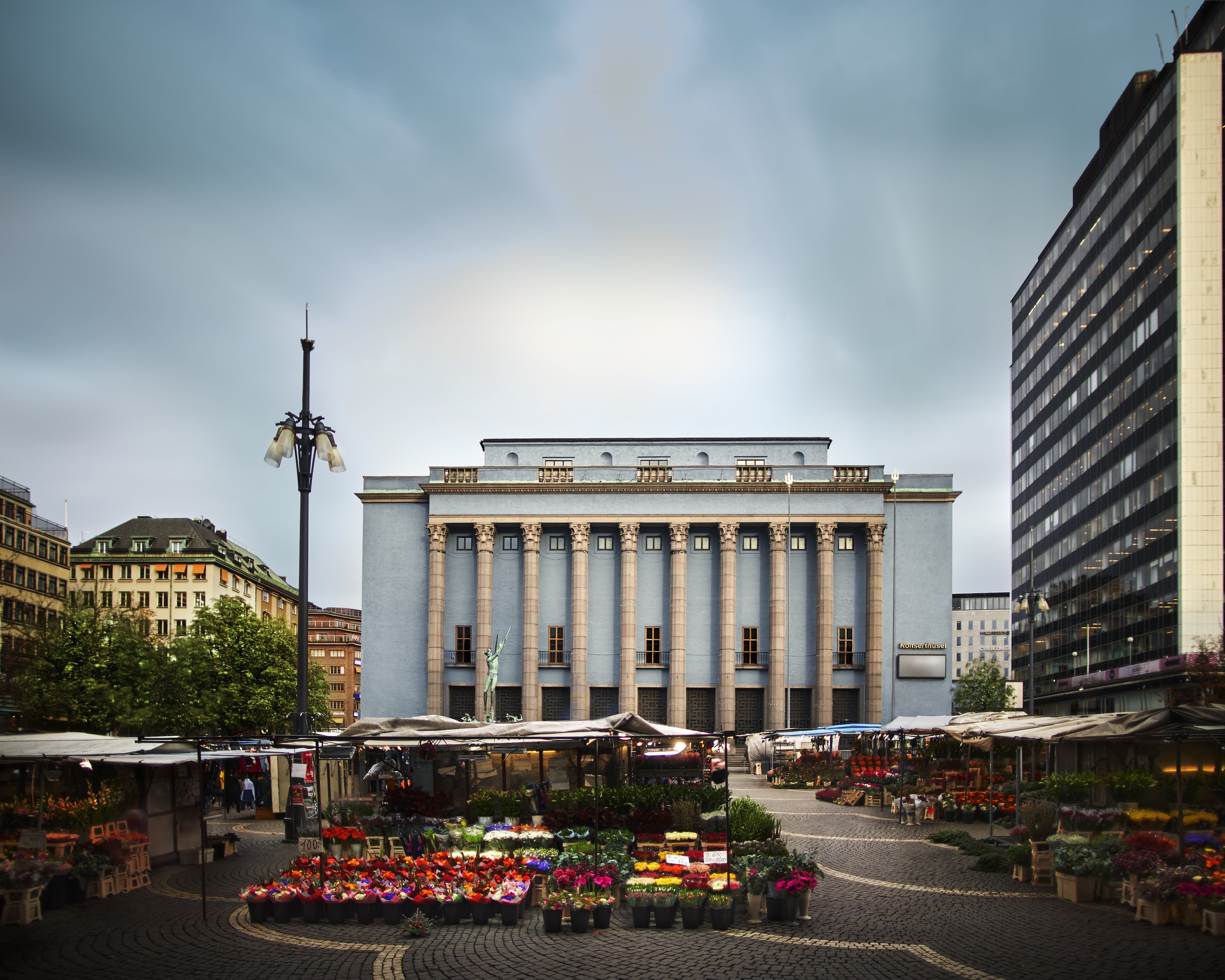 Open-air flower market with various colorful flowers and stalls in a city square, surrounded by buildings, including a large blue, neoclassical building with columns in the background under a cloudy sky.