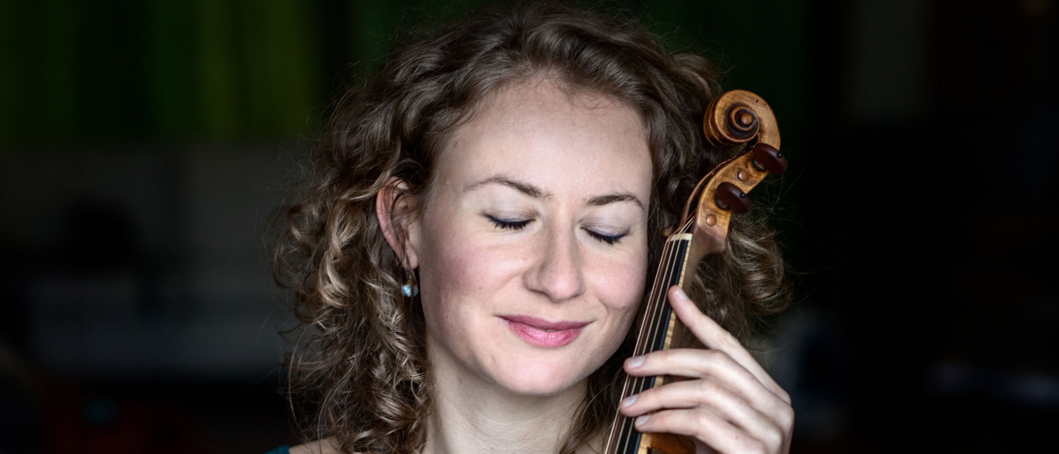 Eine Frau mit lockigem Haar lächelt sanft mit geschlossenen Augen und hält eine Geige an ihre Wange, um einen Moment der friedlichen Verbindung mit ihrem Instrument zu suggerieren.