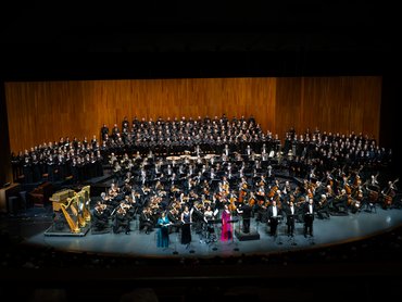 Ein großes Orchester und ein Chor treten auf einer gut beleuchteten, holzgetäfelten Bühne auf. Mehrere Solisten stehen vorne, einer in einem leuchtend rosafarbenen Kleid, während das Publikum aus dem abgedunkelten Theater zuschaut.