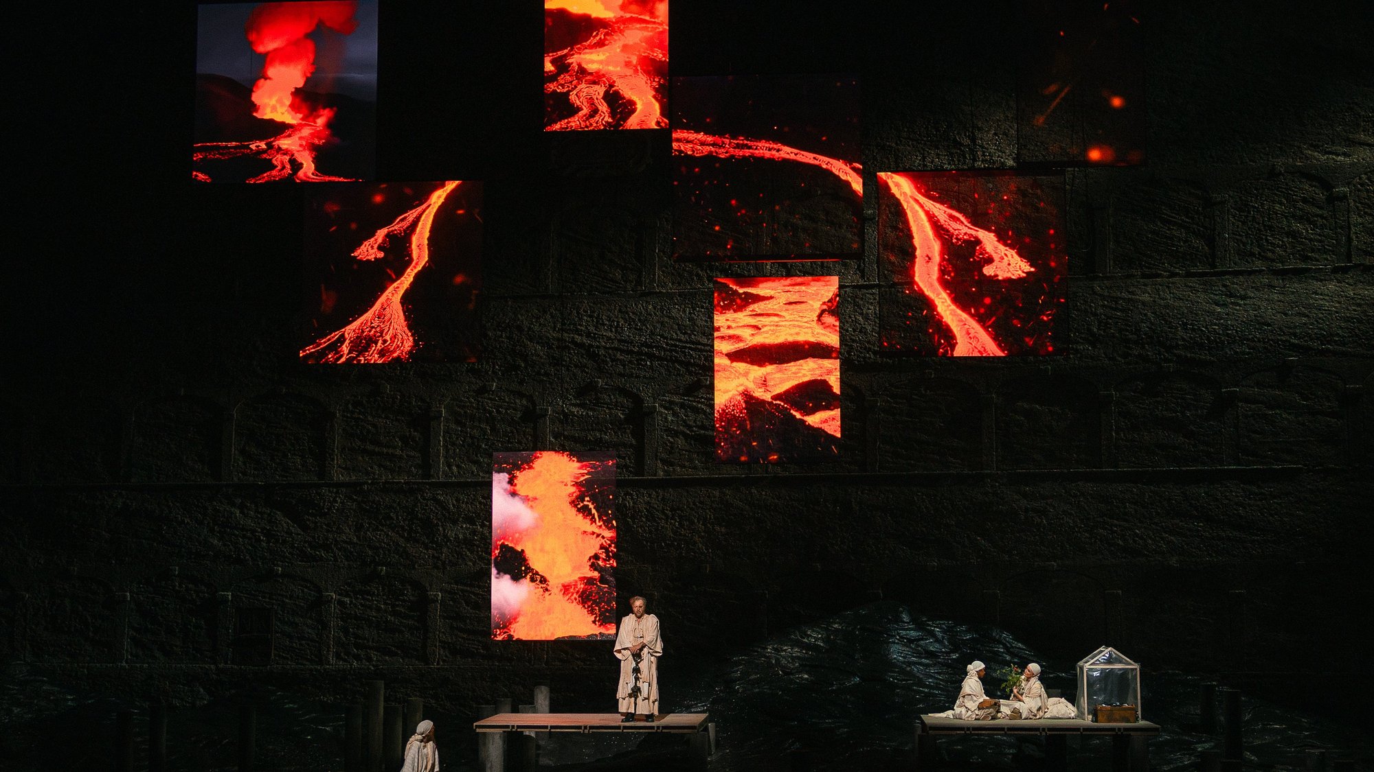 Die Schauspieler treten auf der Bühne unter mehreren großen Bildschirmen auf, die lebendige Bilder von fließender geschmolzener Lava und ausbrechenden Vulkanen zeigen und eine dramatische und intensive Atmosphäre schaffen.