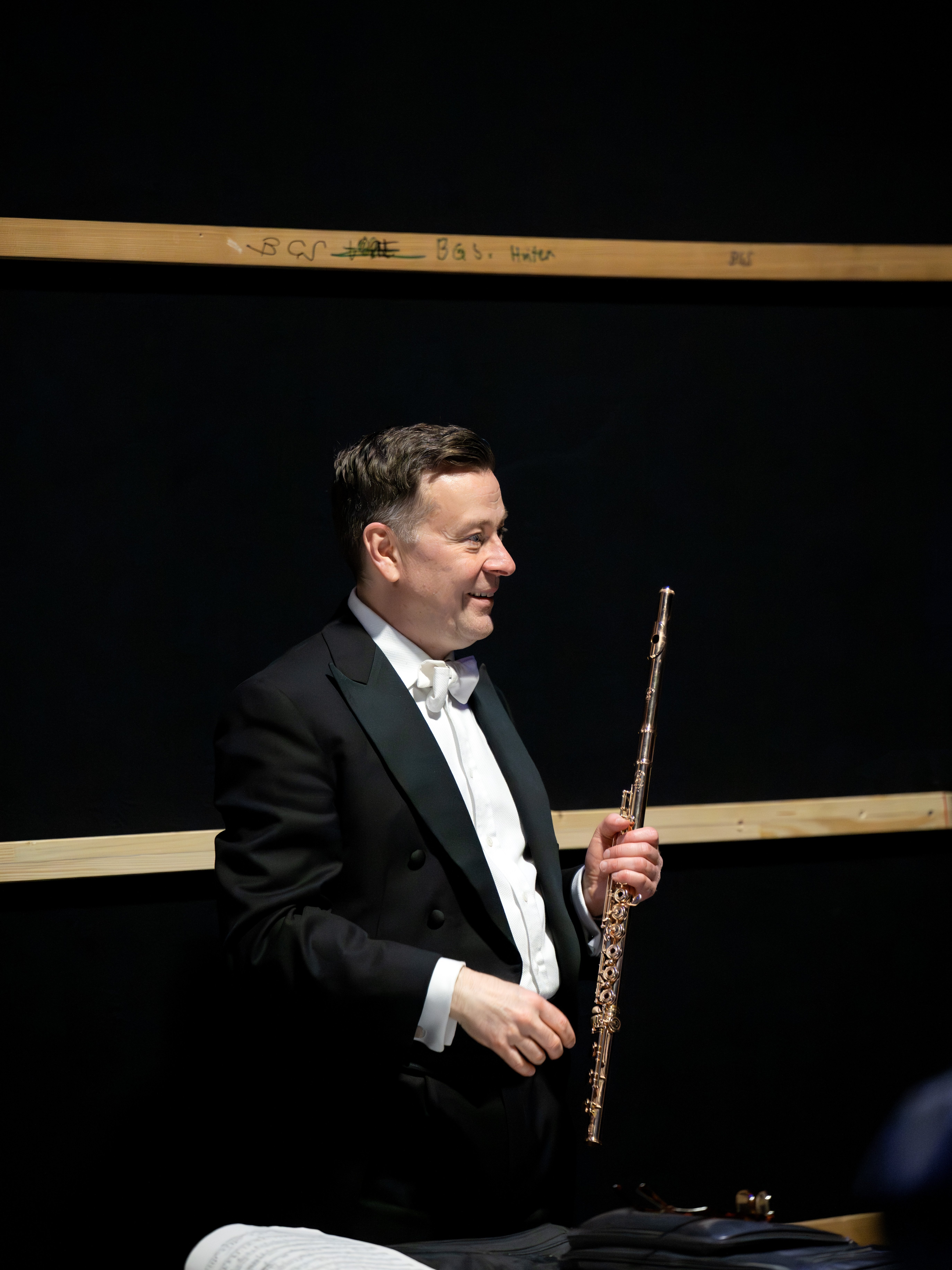 Ein Mann im schwarzen Smoking und mit Fliege steht in einem Haus und hält eine Flöte. Er lächelt und hat einen dunklen Hintergrund mit Holzbalken hinter sich.