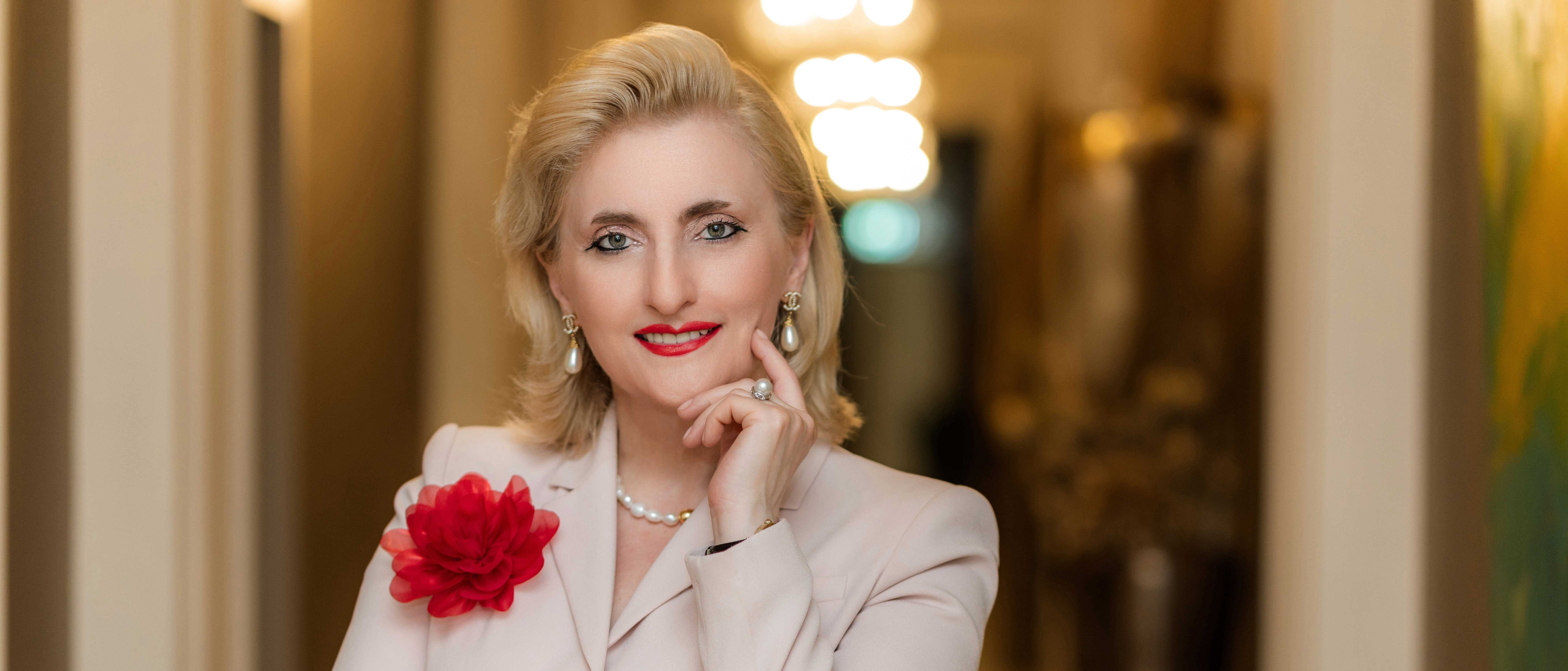 Eine Frau mit blonden Haaren, die einen cremefarbenen Anzug, Perlenschmuck und roten Lippenstift trägt, steht in einem Raum und lächelt. Sie hat eine rote Blume am Revers und stützt nachdenklich die Hand am Kinn.