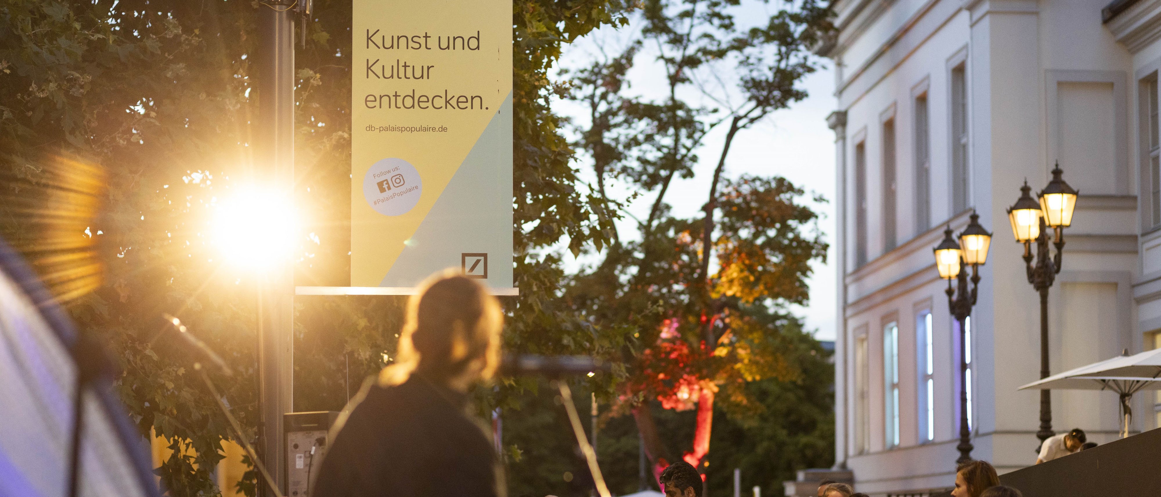 Ein Banner für das PalaisPopulaire, das an einem Mast über einer Menschenmenge hängt und auf dem Kunst und Kultur entdecken steht.