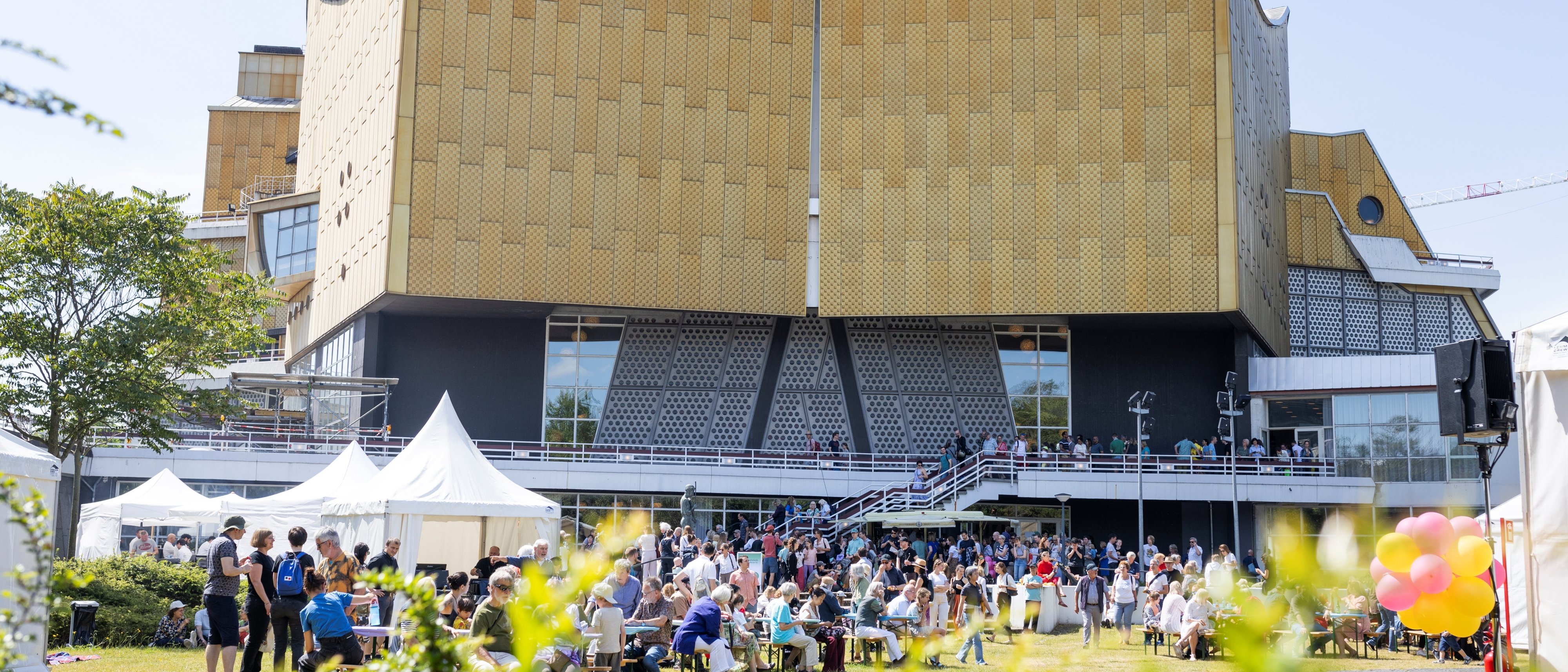 A large crowd gathers in front of a modern, gold-coloured building, the Philharmonie Berlin, with a unique architectural design. On this sunny day, people sit and stand, tents and colourful balloons add to the festive atmosphere.