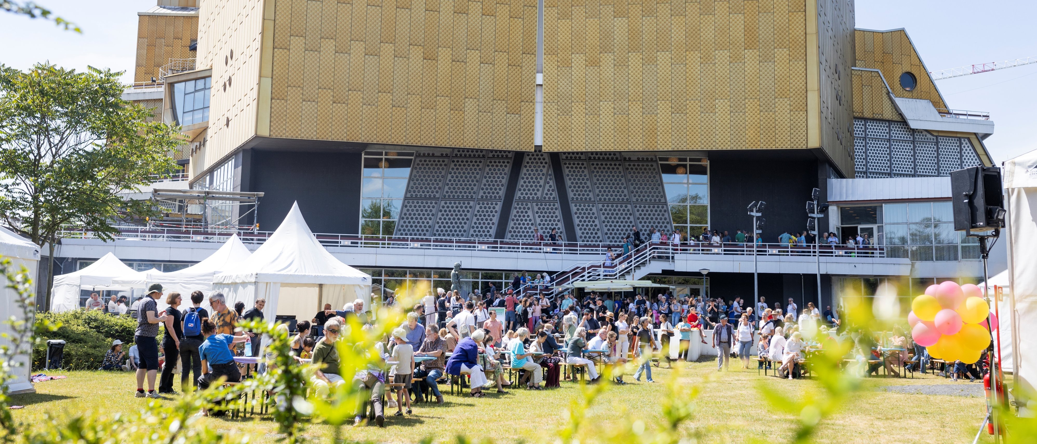 Eine große Menschenmenge versammelt sich vor einem modernen, goldfarbenen Gebäude mit einzigartigem architektonischen Design. An diesem sonnigen Tag sitzen und stehen die Menschen, Zelte und bunte Luftballons tragen zur festlichen Atmosphäre bei.