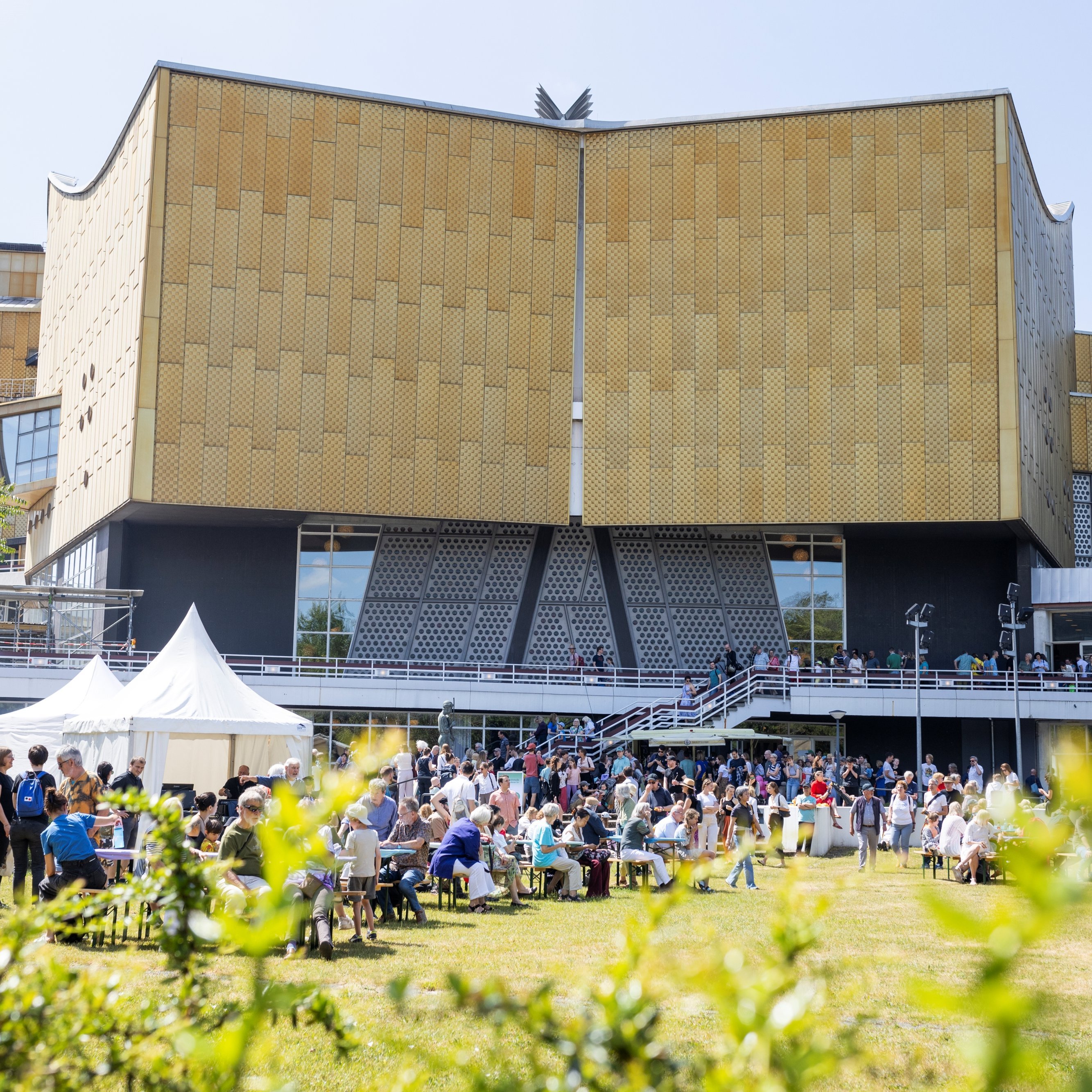 Eine große Menschenmenge versammelt sich vor einem modernen, goldfarbenen Gebäude mit einzigartigem architektonischen Design. An diesem sonnigen Tag sitzen und stehen die Menschen, Zelte und bunte Luftballons tragen zur festlichen Atmosphäre bei.