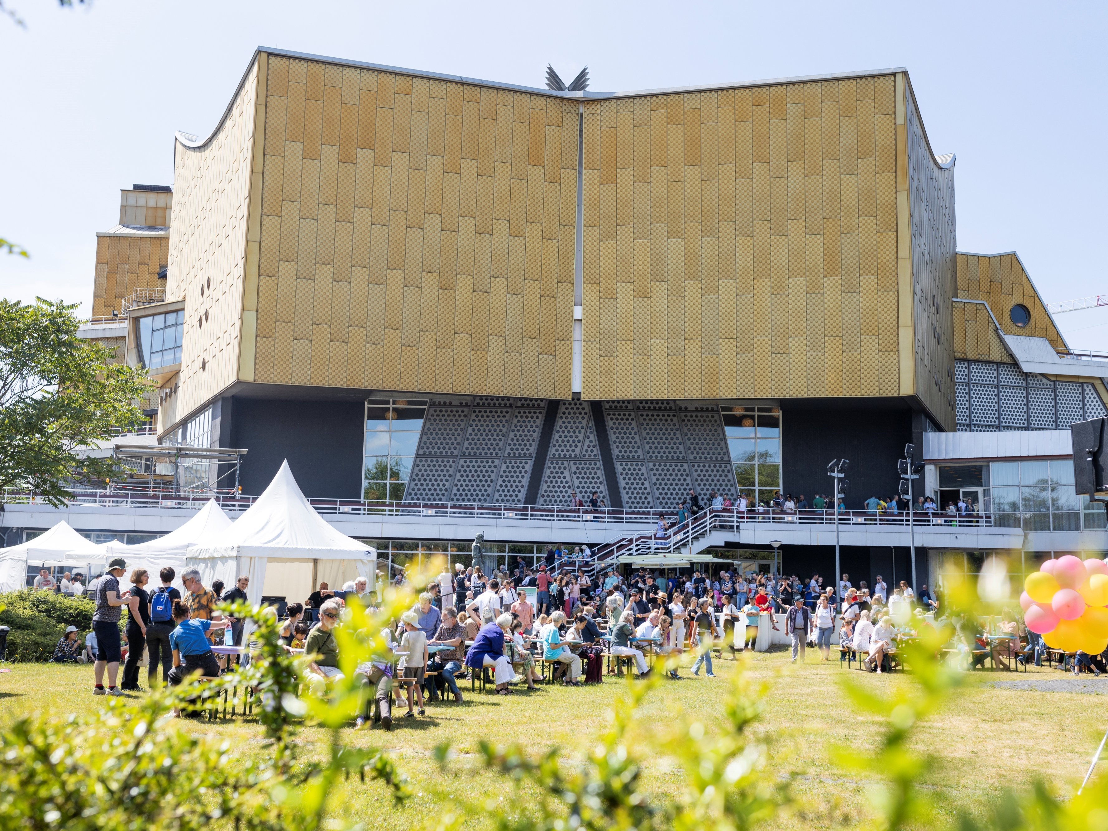 A large crowd gathers in front of a modern, gold-coloured building, the Philharmonie Berlin, with a unique architectural design. On this sunny day, people sit and stand, tents and colourful balloons add to the festive atmosphere.