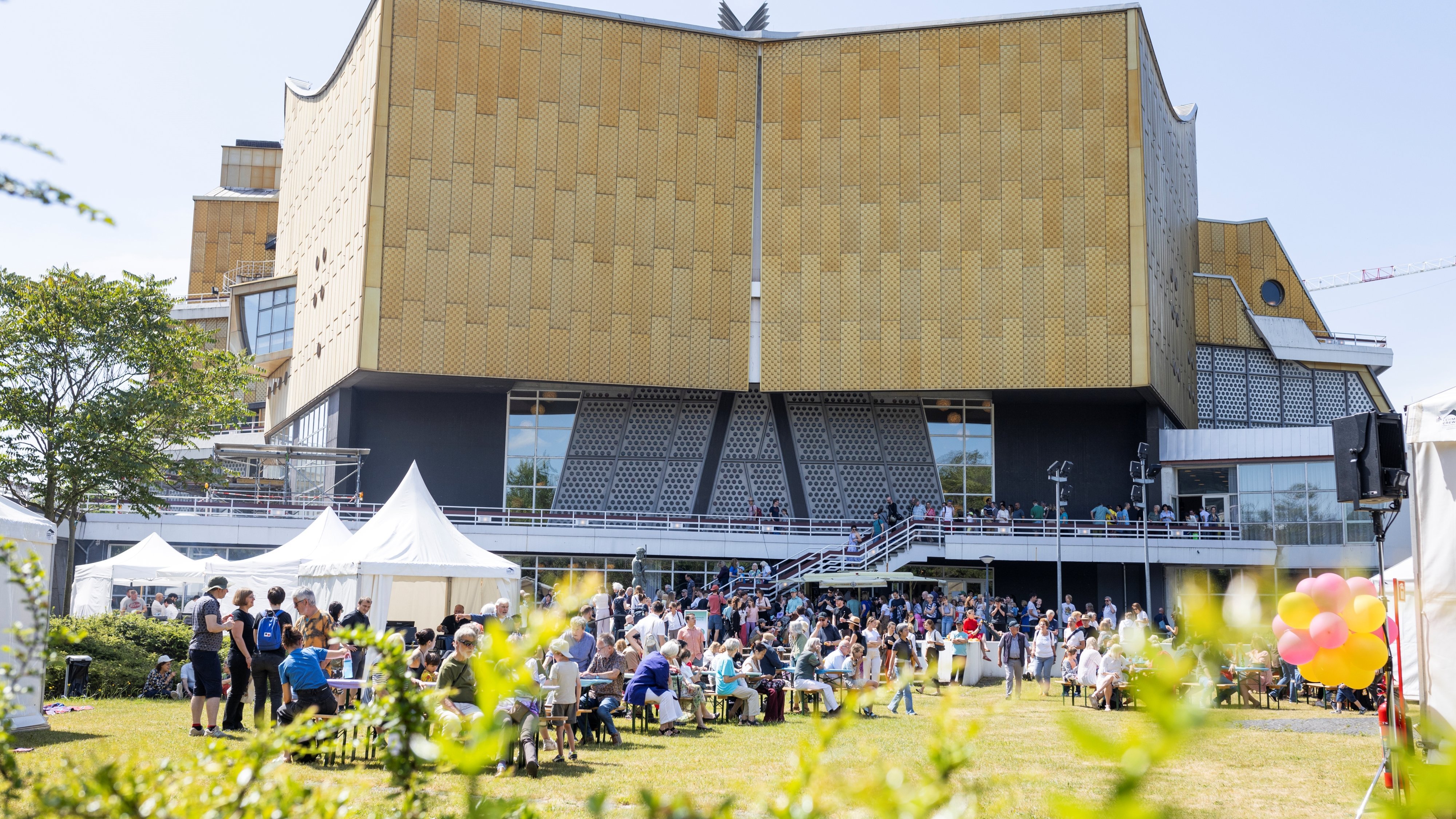 A large crowd gathers in front of a modern, gold-coloured building, the Philharmonie Berlin, with a unique architectural design. On this sunny day, people sit and stand, tents and colourful balloons add to the festive atmosphere.
