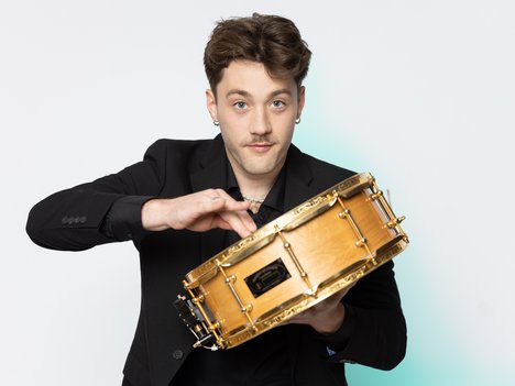A young man with short brown hair and a mustache holds a snare drum and appears to tap it with one hand. He wears a black jacket and stands against a plain white background.