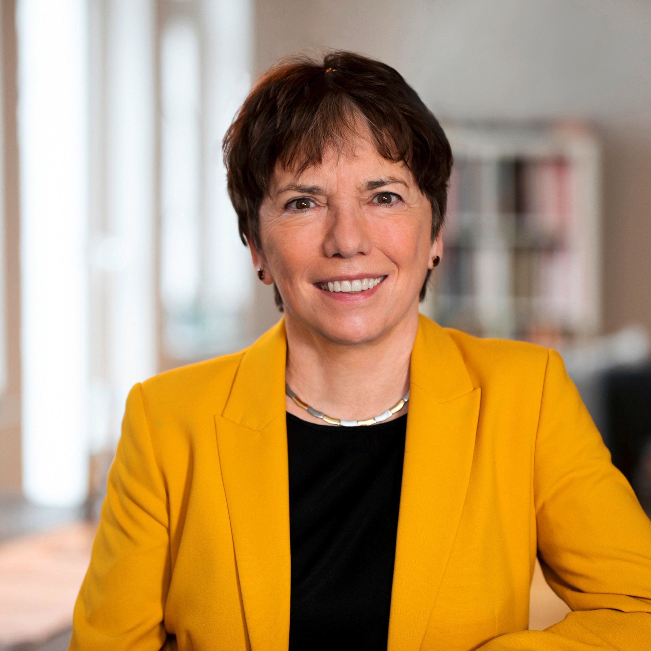 Eine Frau mit kurzen braunen Haaren, die einen gelben Blazer über einem schwarzen Oberteil trägt, lächelt in einem hellen, modernen Raum mit weichem natürlichem Licht in die Kamera.