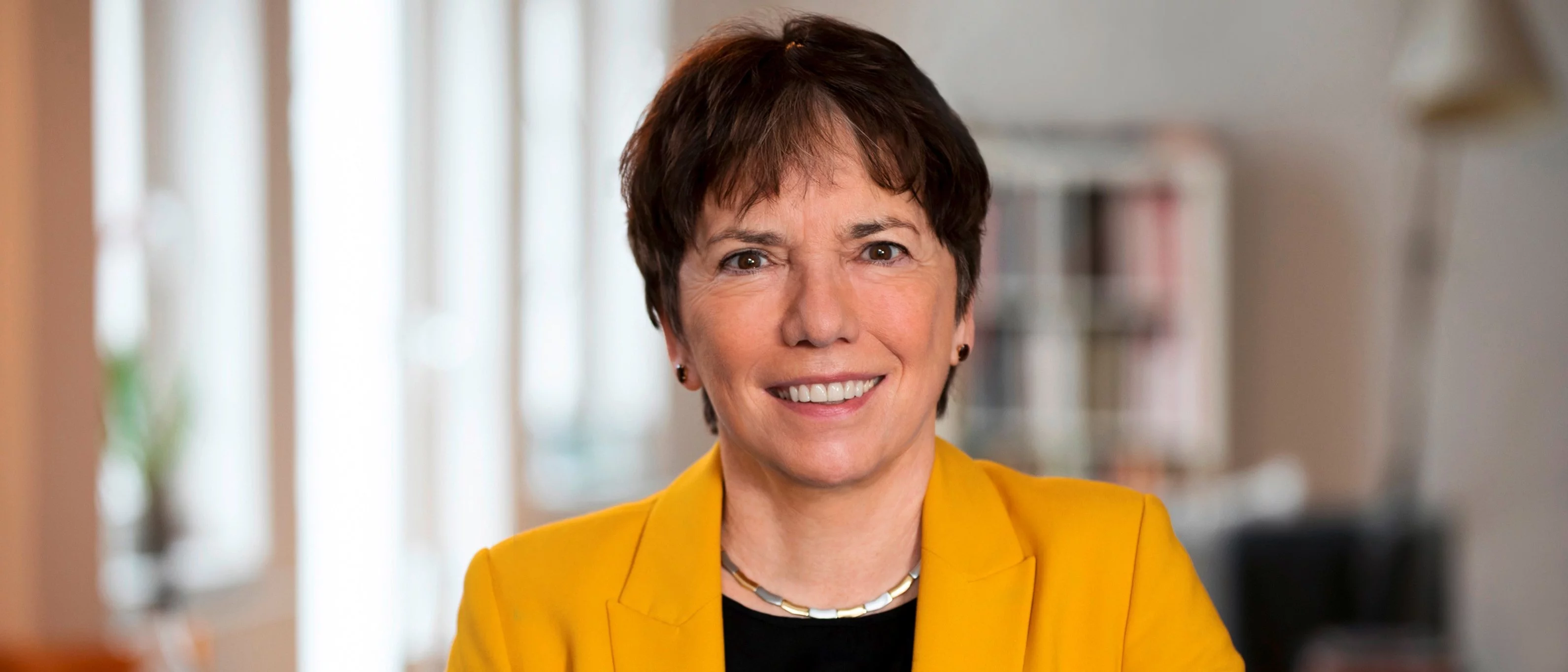 A woman with short brown hair, wearing a yellow blazer and black top, smiles at the camera while sitting indoors in a brightly lit room with blurred background.