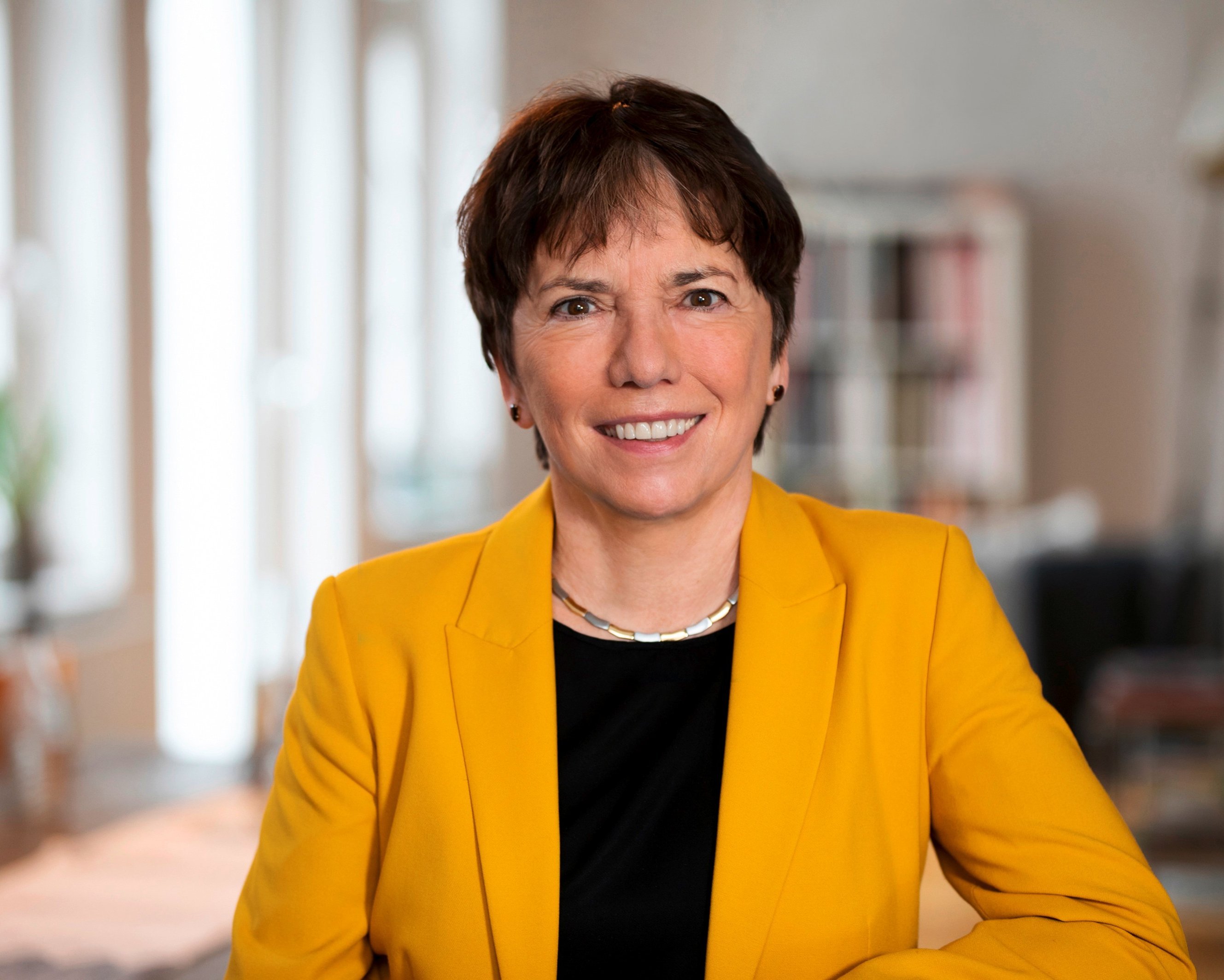 Eine Frau mit kurzen braunen Haaren, die einen gelben Blazer über einem schwarzen Oberteil trägt, lächelt in einem hellen, modernen Raum mit weichem natürlichem Licht in die Kamera.
