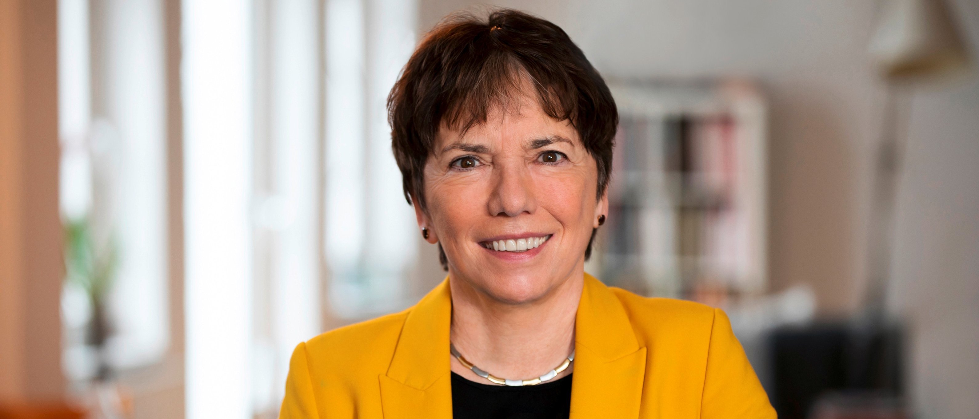 Eine Frau mit kurzen braunen Haaren, die einen gelben Blazer über einem schwarzen Oberteil trägt, lächelt in einem hellen, modernen Raum mit weichem natürlichem Licht in die Kamera.