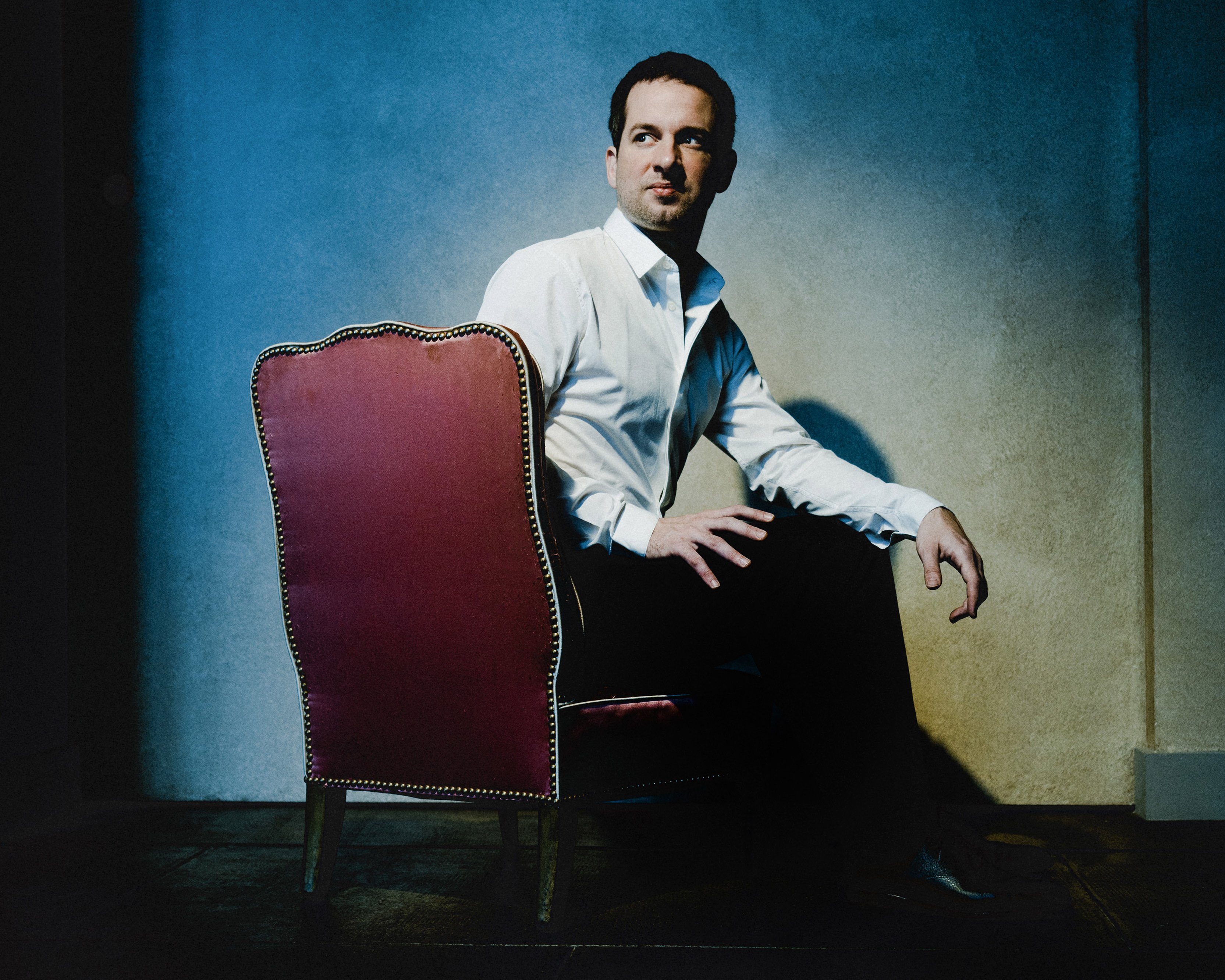 Bertrand Chamayou in einem weißen Hemd sitzt seitlich auf einem roten Stuhl, dreht den Kopf zur Seite und blickt in einen schwach beleuchteten Raum mit strukturierten Wänden und dramatischer Beleuchtung.