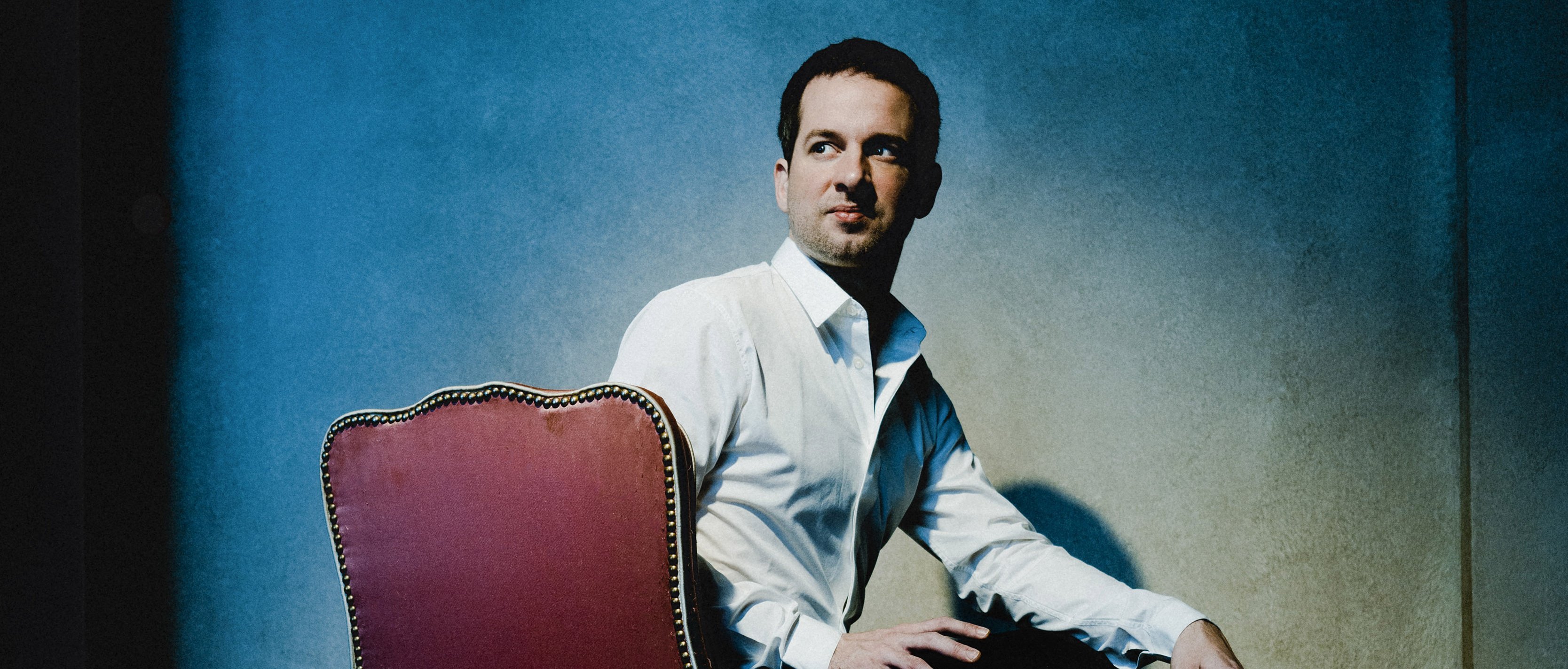 Bertrand Chamayou in einem weißen Hemd sitzt seitlich auf einem roten Stuhl, dreht den Kopf zur Seite und blickt in einen schwach beleuchteten Raum mit strukturierten Wänden und dramatischer Beleuchtung.