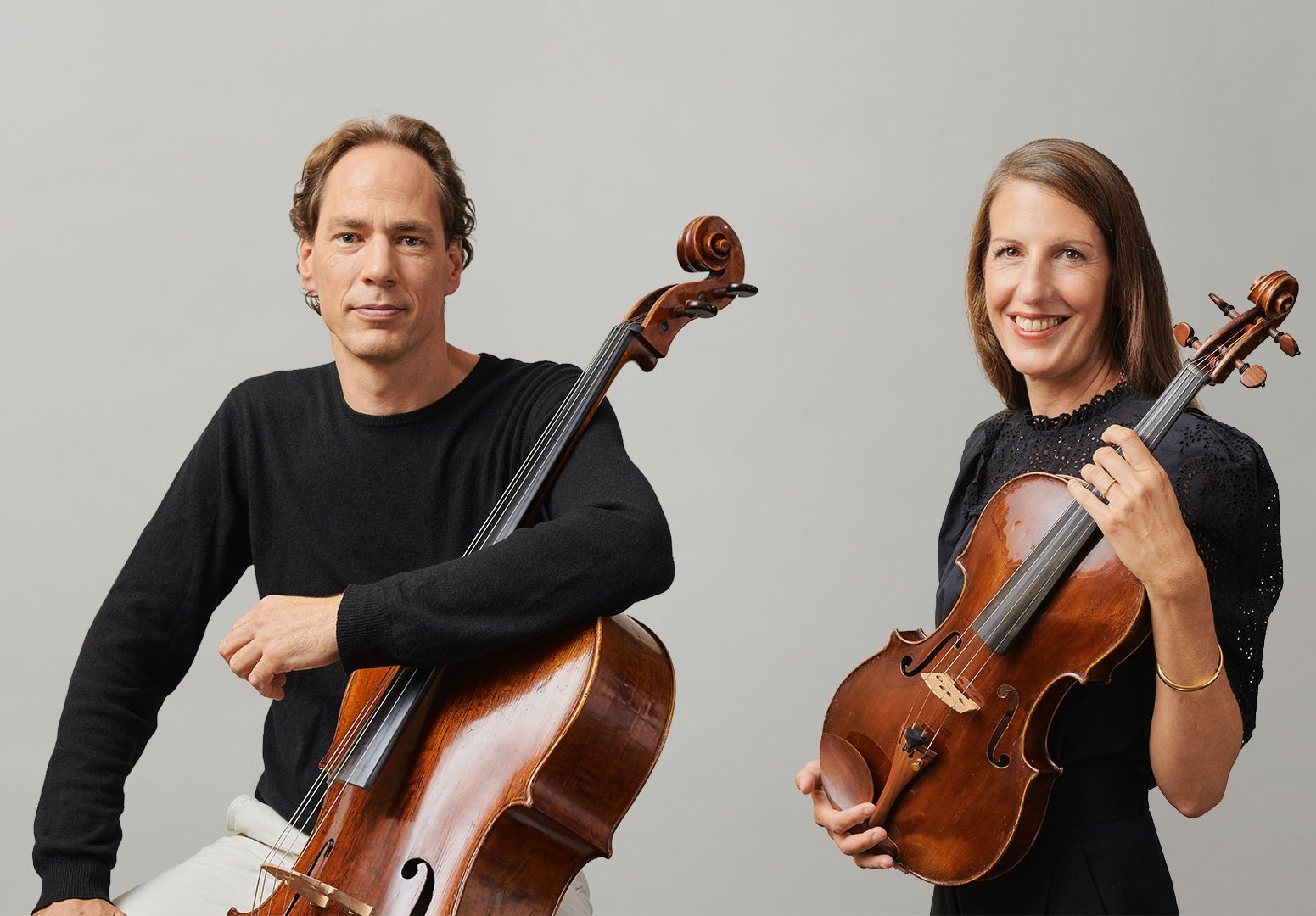 Ein Mann mit einem Cello und eine Frau mit einer Bratsche sitzen Seite an Seite vor einem schlichten hellgrauen Hintergrund, beide lächeln und tragen schwarze Oberteile.