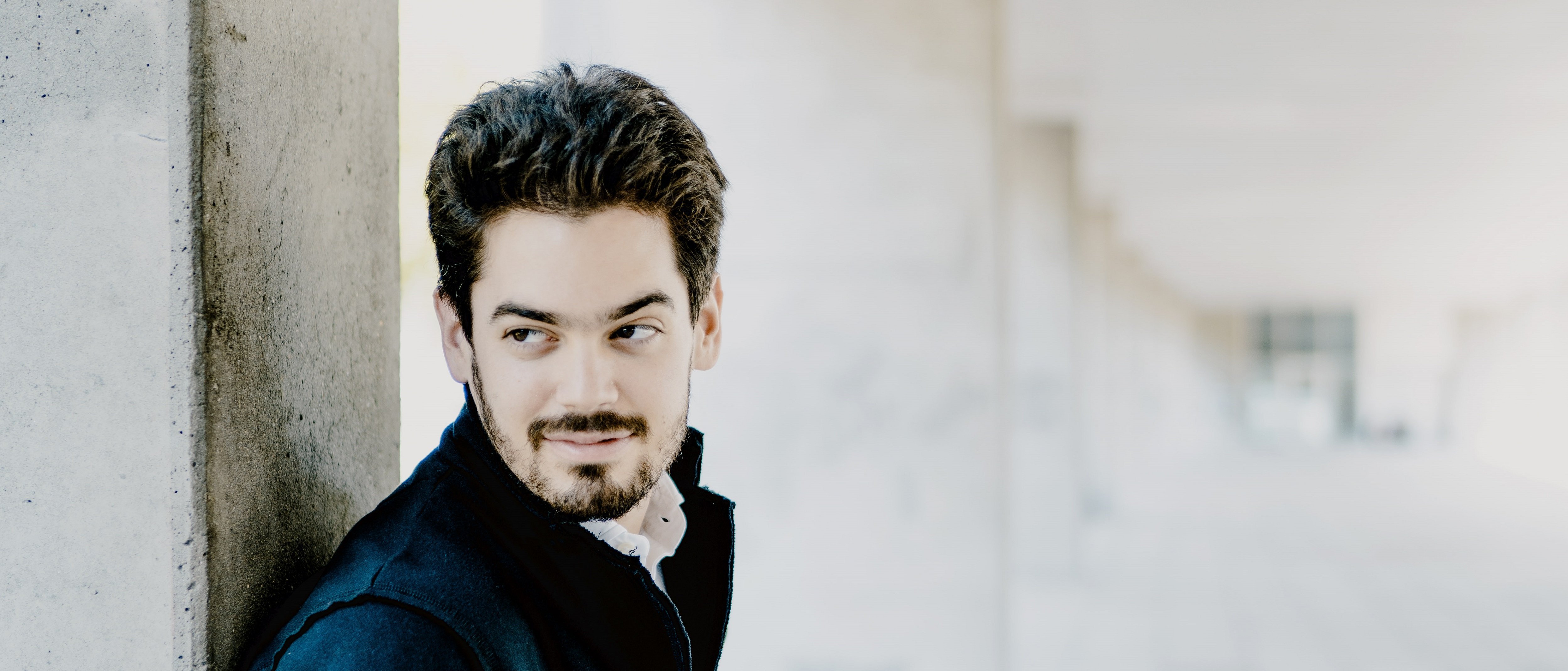 Das Bild zeigt Lahav Shani mit Bart und dunklem Haar, der in einem Flur an einem Betonpfeiler lehnt. Er trägt eine dunkle Jacke und Hose und blickt nachdenklich zur Seite. Die Szenerie umfasst einen gefliesten Boden und eine Decke mit modernen Designelementen.