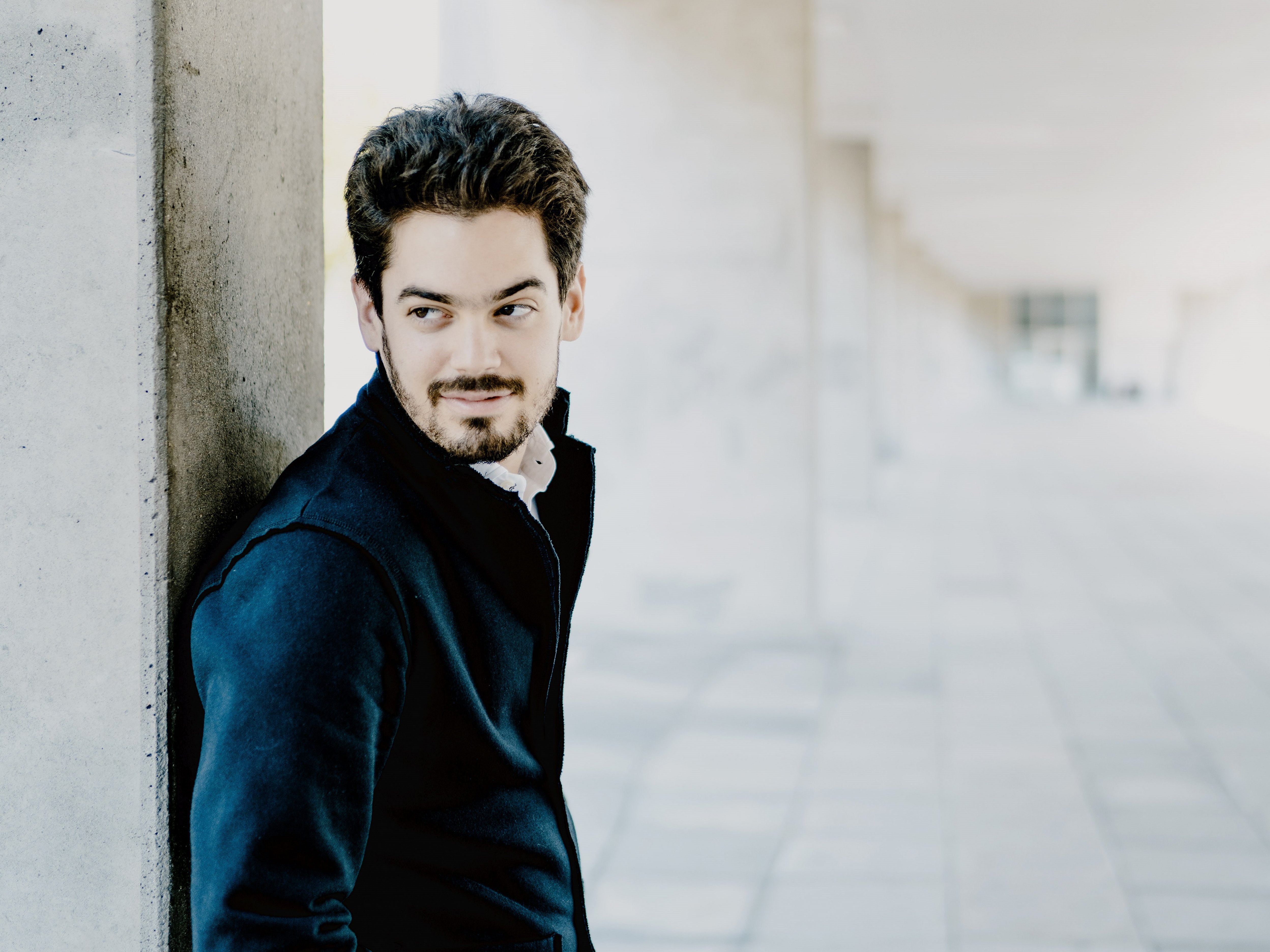 Das Bild zeigt Lahav Shani mit Bart und dunklem Haar, der in einem Flur an einem Betonpfeiler lehnt. Er trägt eine dunkle Jacke und Hose und blickt nachdenklich zur Seite. Die Szenerie umfasst einen gefliesten Boden und eine Decke mit modernen Designelementen.