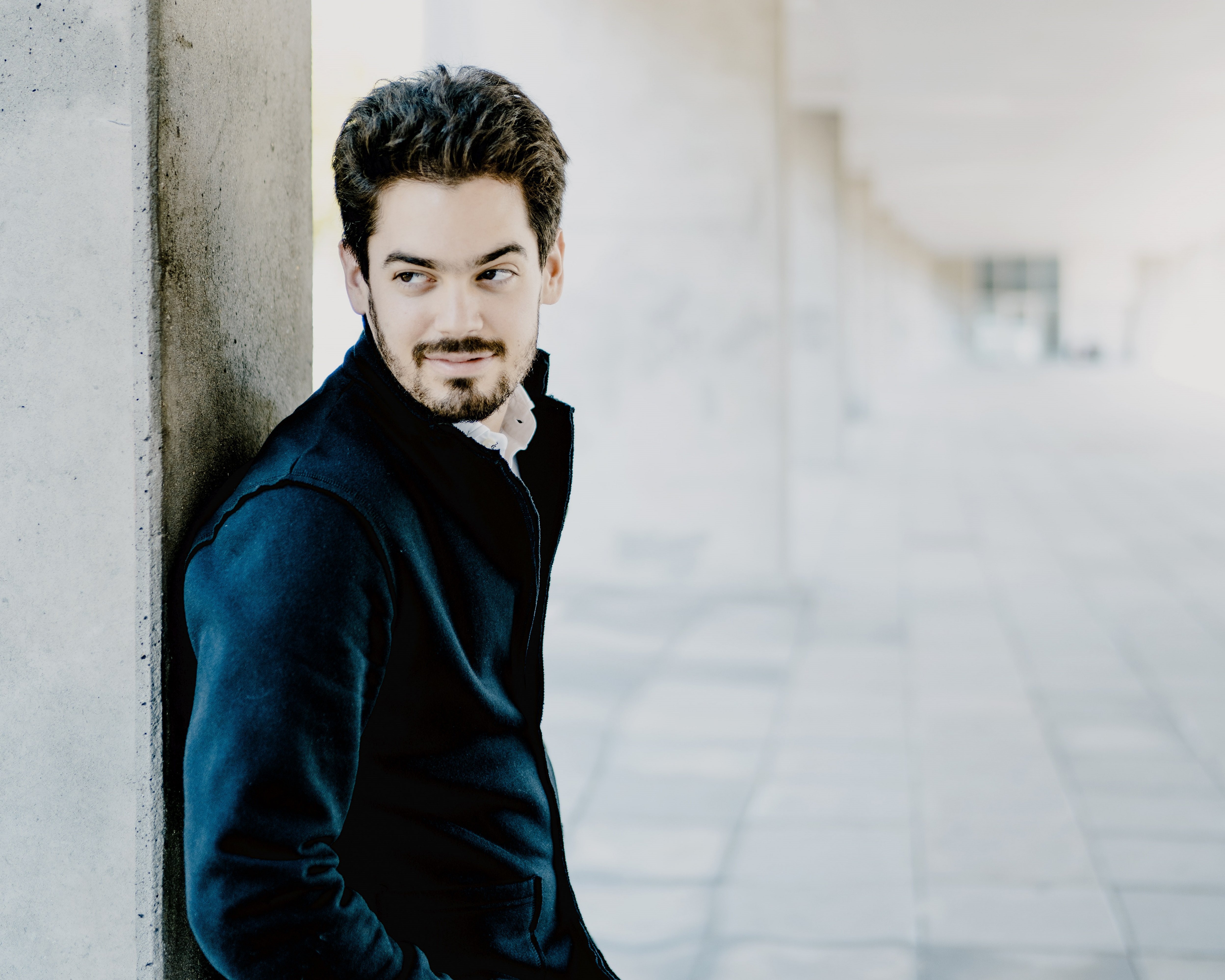 Das Bild zeigt Lahav Shani mit Bart und dunklem Haar, der in einem Flur an einem Betonpfeiler lehnt. Er trägt eine dunkle Jacke und Hose und blickt nachdenklich zur Seite. Die Szenerie umfasst einen gefliesten Boden und eine Decke mit modernen Designelementen.