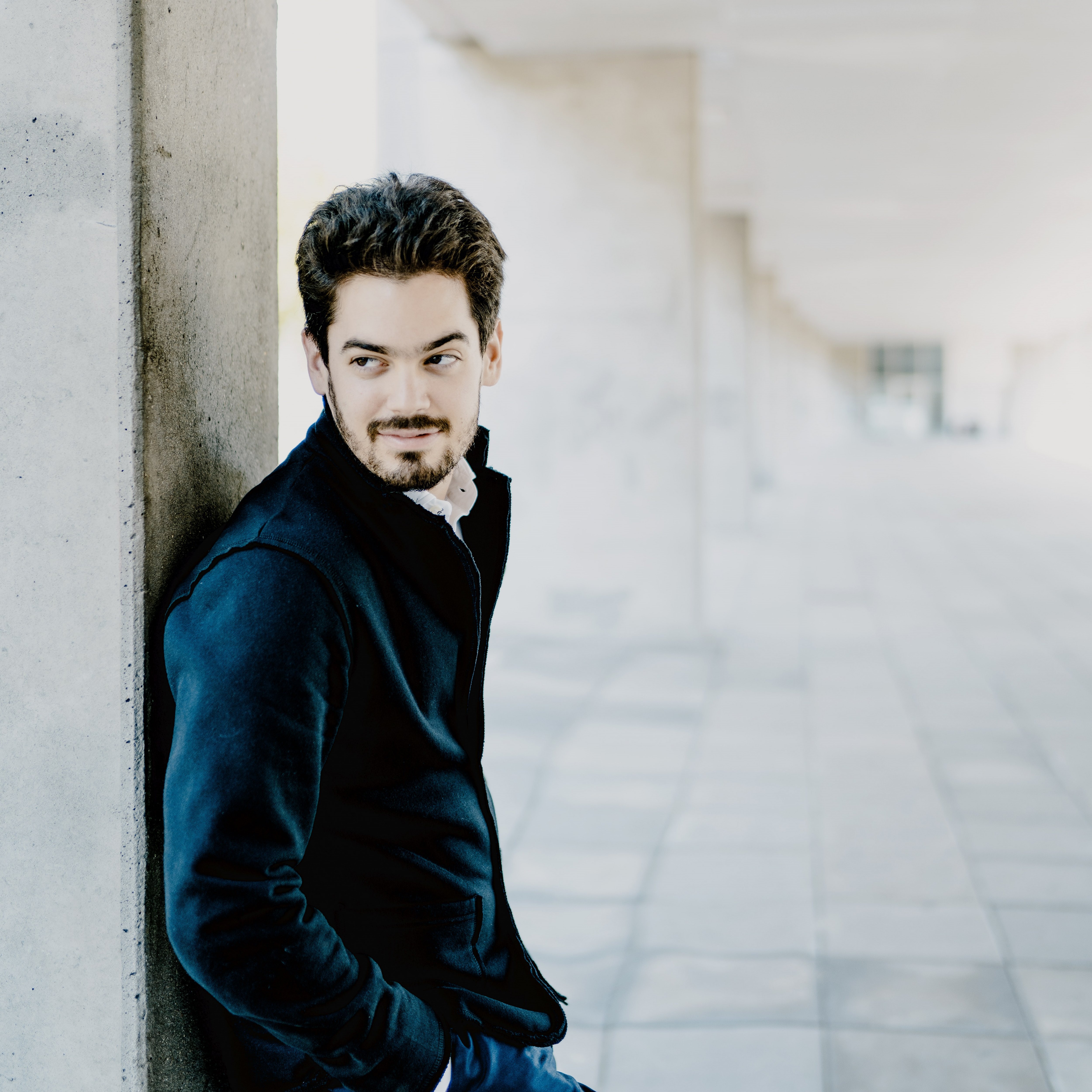 Das Bild zeigt Lahav Shani mit Bart und dunklem Haar, der in einem Flur an einem Betonpfeiler lehnt. Er trägt eine dunkle Jacke und Hose und blickt nachdenklich zur Seite. Die Szenerie umfasst einen gefliesten Boden und eine Decke mit modernen Designelementen.