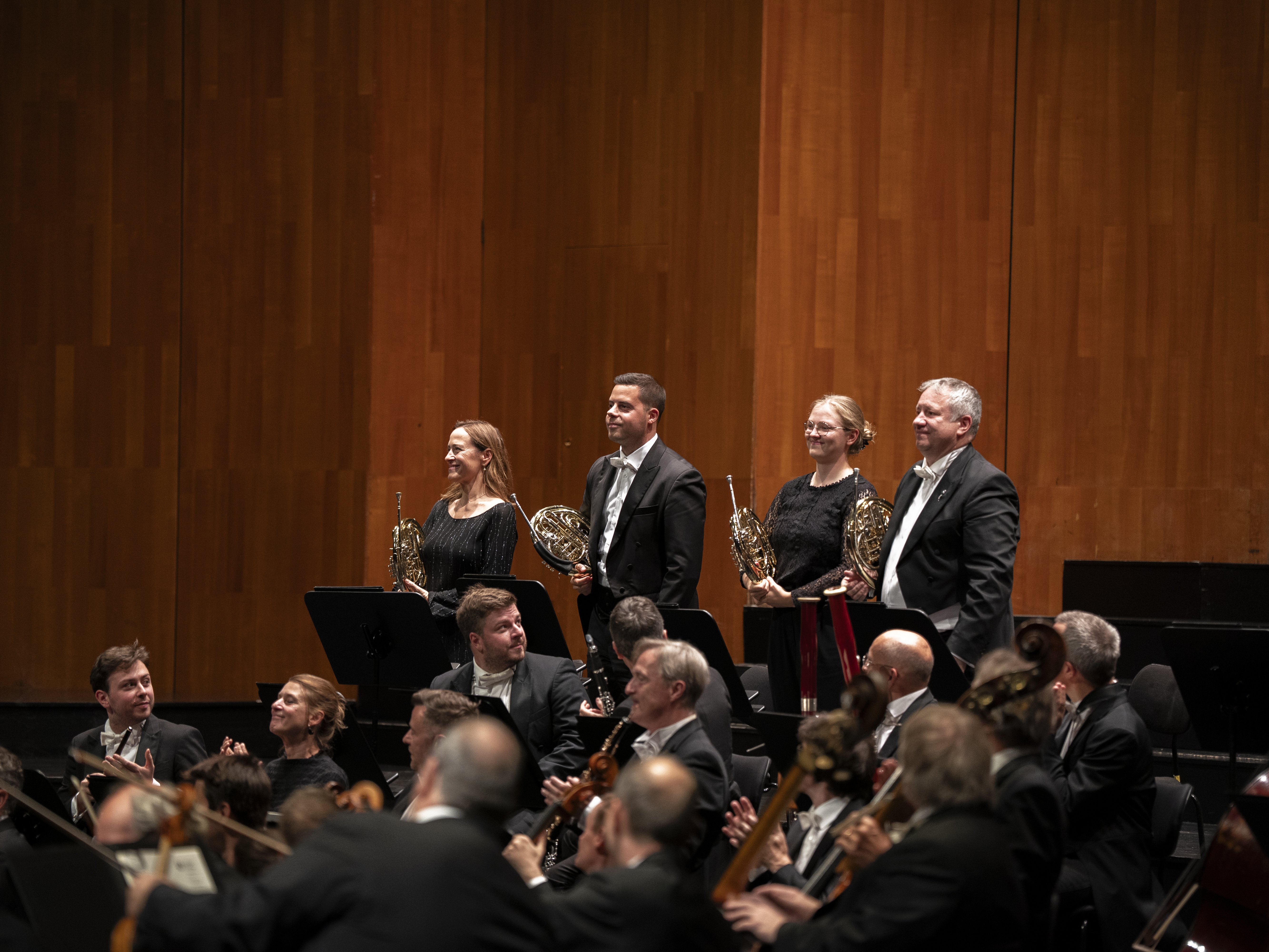 Vier Hornist*innen stehen lächelnd mit ihren Instrumenten in der Hand da, während der Rest des Orchesters, das mit Streichinstrumenten besetzt ist, zu ihnen hinblickt. Im Hintergrund ist eine Holzwand eines Konzertsaals zu sehen.