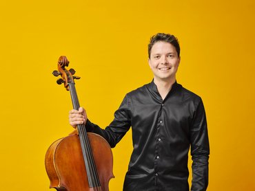 A smiling Moritz Huemer in a black shirt stands against a yellow background, holding a cello upright with one hand.