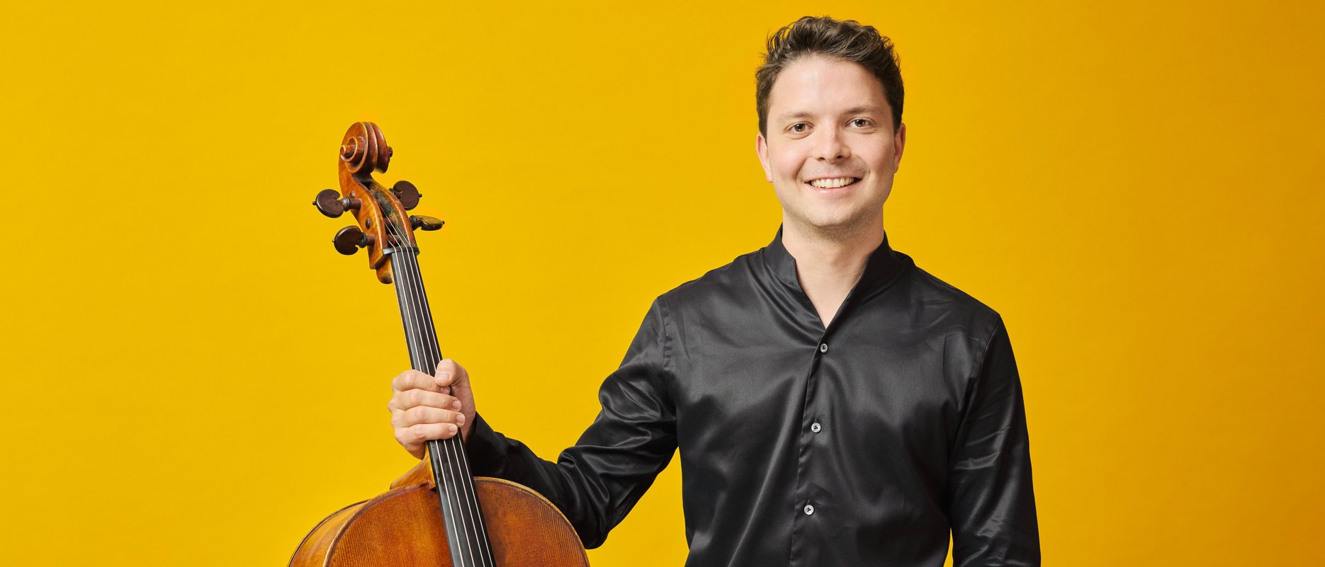 A smiling Moritz Huemer in a black shirt stands against a yellow background, holding a cello upright with one hand.