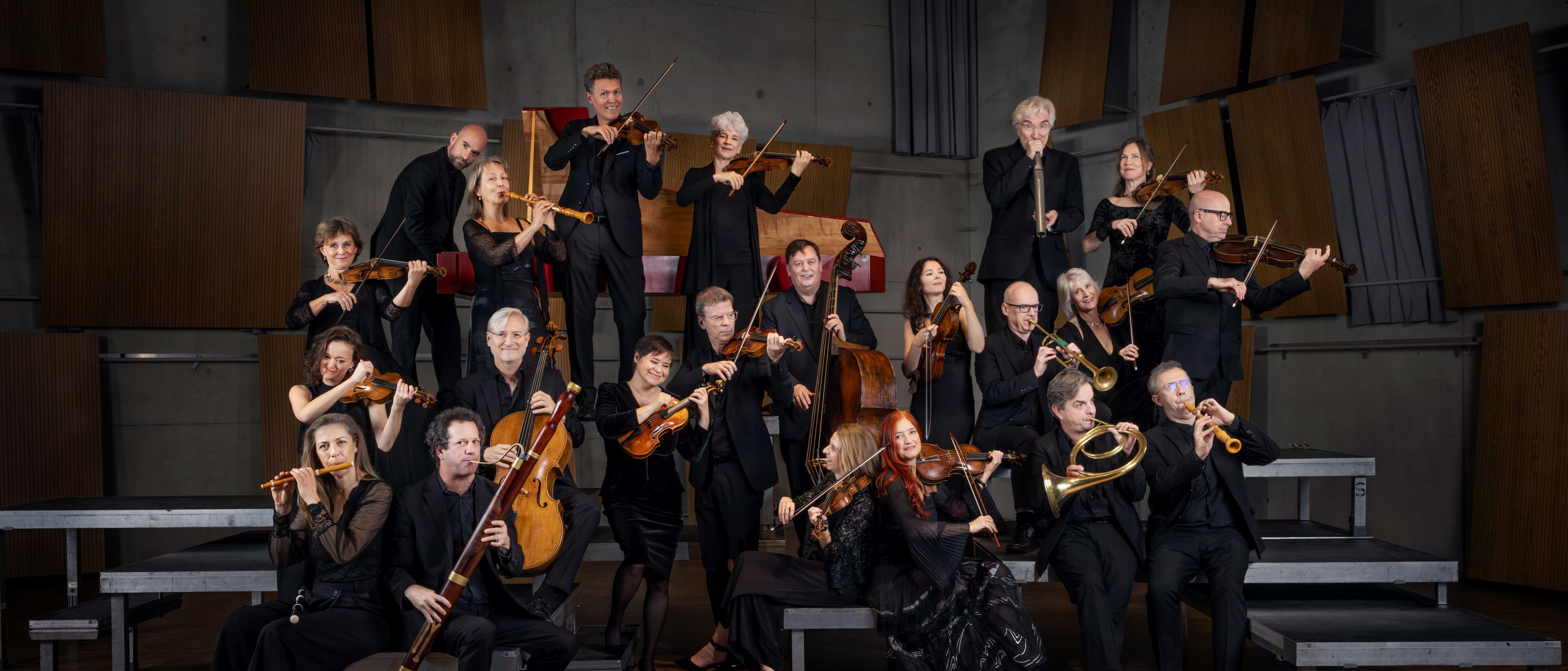 Eine Gruppe von schwarz gekleideten Musikern posiert mit ihren Instrumenten, darunter Geigen, Celli, Trompeten und Holzblasinstrumente, auf gestuften Podesten in einem holzgetäfelten Konzertsaal.