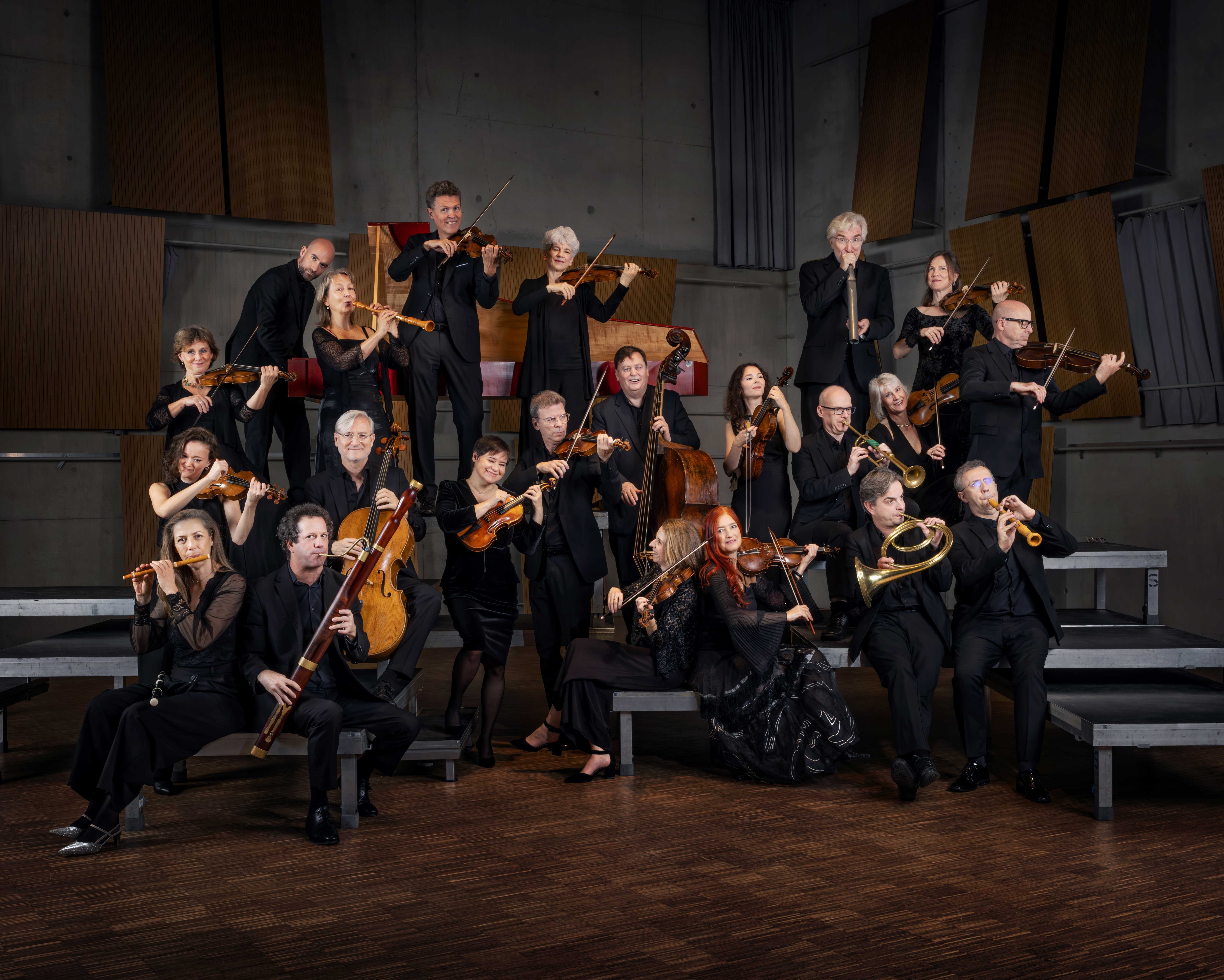Eine Gruppe von schwarz gekleideten Musikern posiert mit ihren Instrumenten, darunter Geigen, Celli, Trompeten und Holzblasinstrumente, auf gestuften Podesten in einem holzgetäfelten Konzertsaal.