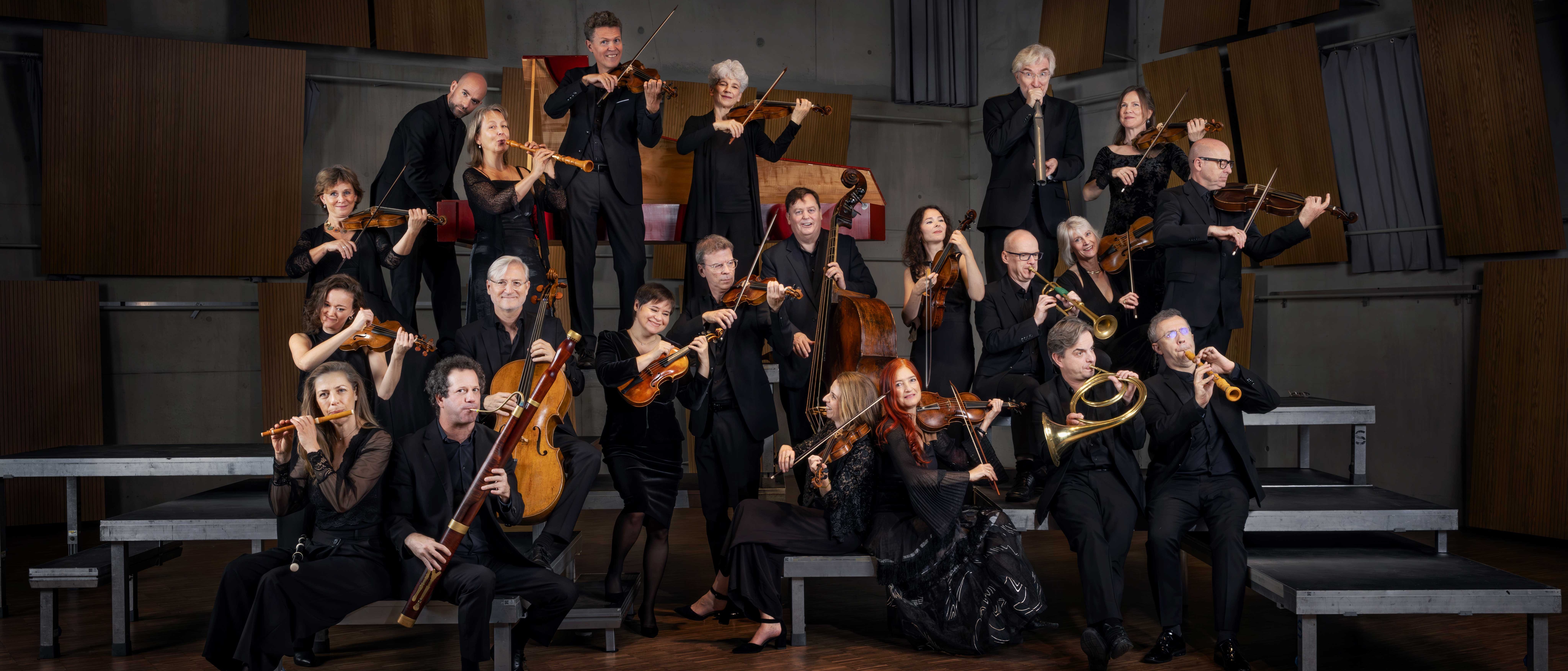 A group of formally dressed musicians pose with their string and brass instruments on tiered platforms in a modern, wood-paneled room, smiling and looking at the camera.