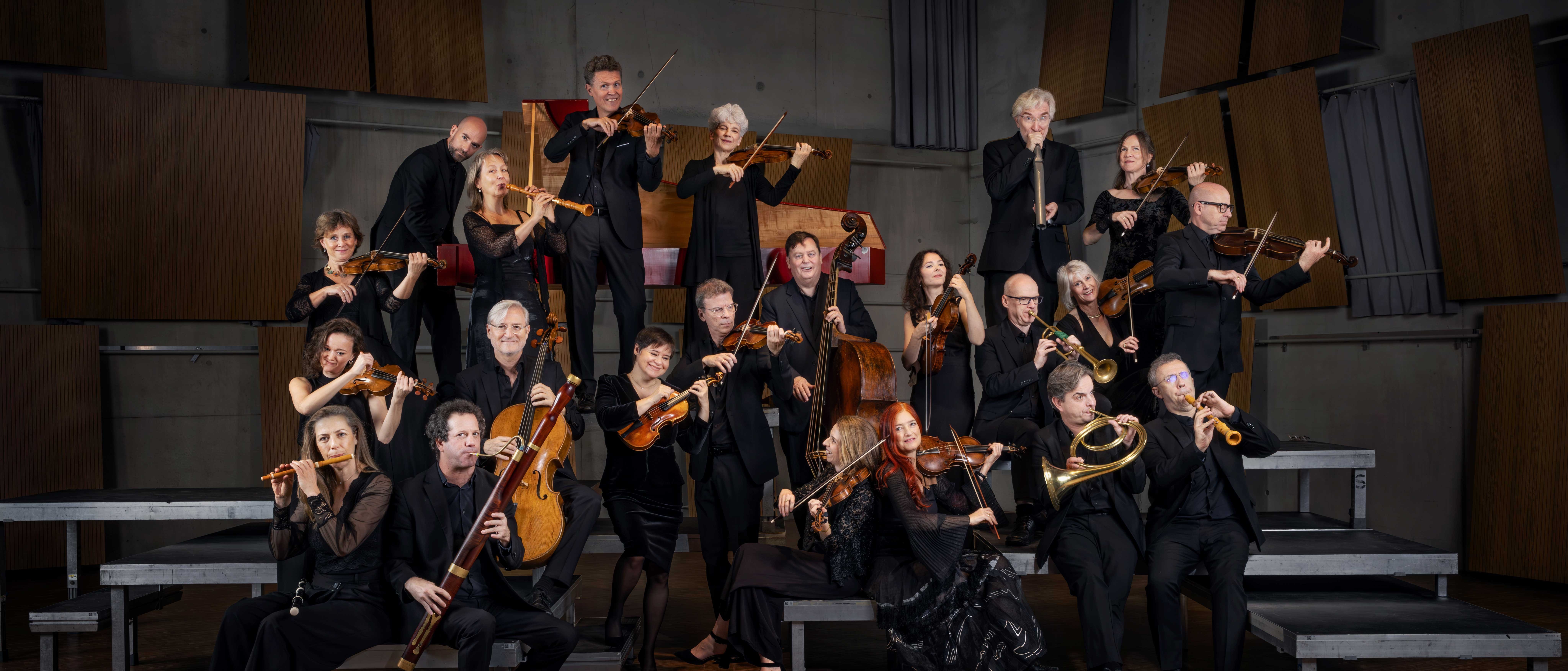 Eine Gruppe von schwarz gekleideten Musikern posiert mit ihren Instrumenten, darunter Geigen, Celli, Trompeten und Holzblasinstrumente, auf gestuften Podesten in einem holzgetäfelten Konzertsaal.