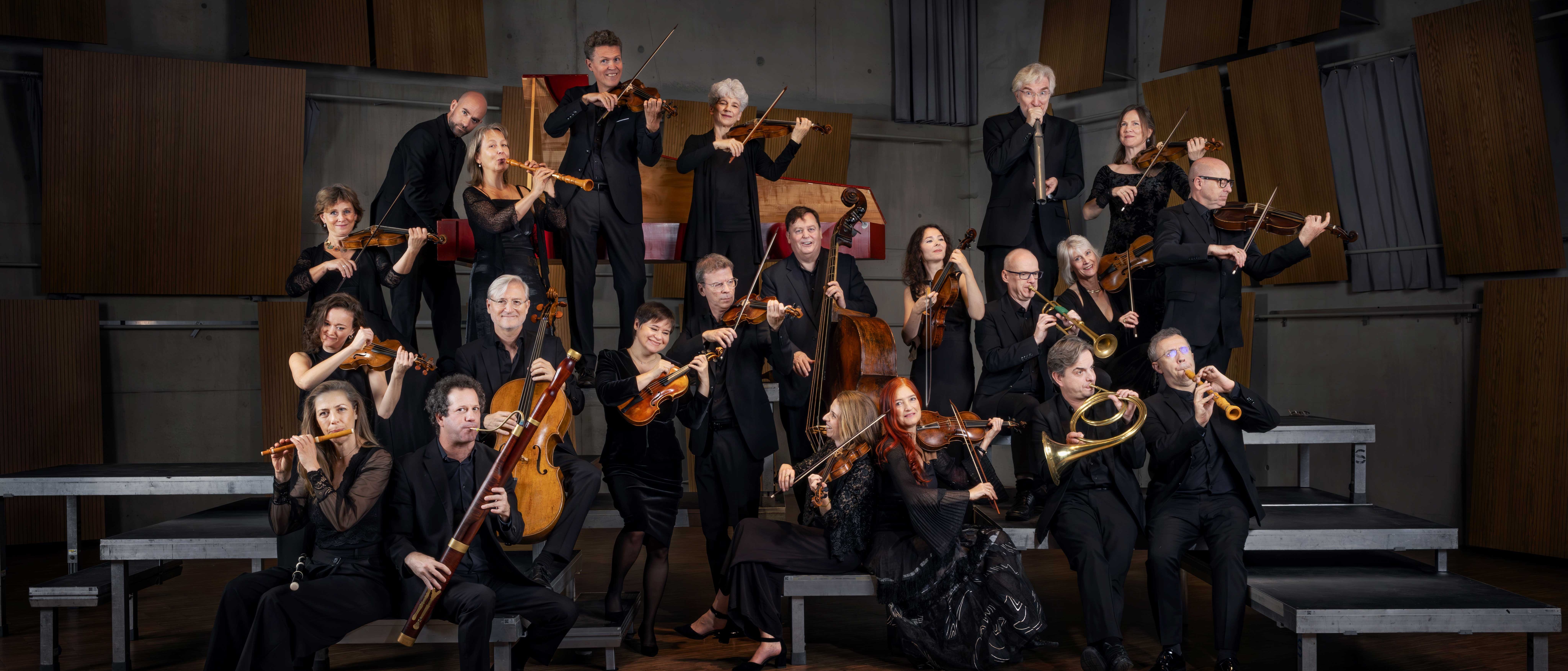 Eine Gruppe von schwarz gekleideten Musikern posiert mit ihren Instrumenten, darunter Geigen, Celli, Trompeten und Holzblasinstrumente, auf gestuften Podesten in einem holzgetäfelten Konzertsaal.