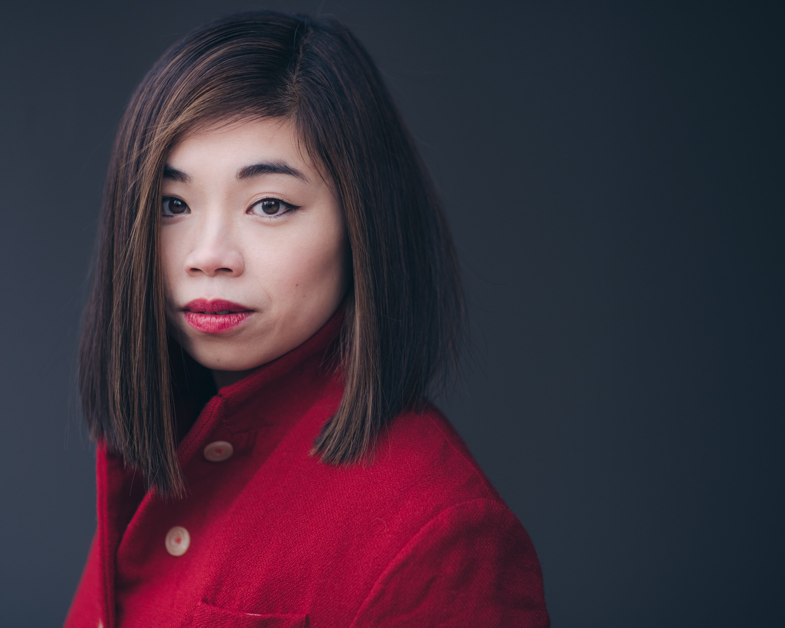 Elim Chan mit glattem, schulterlangem dunklem Haar und rotem Lippenstift trägt einen roten Mantel und blickt vor einem schlichten dunklen Hintergrund direkt in die Kamera.