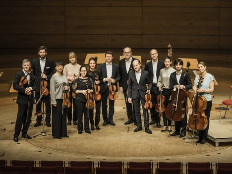 Zwölf Musiker in förmlicher Kleidung stehen auf einer hölzernen Bühne in einem Konzertsaal und halten Streichinstrumente, darunter Geigen, Bratschen und Celli, vor leeren Notenständern und Stühlen im Hintergrund.