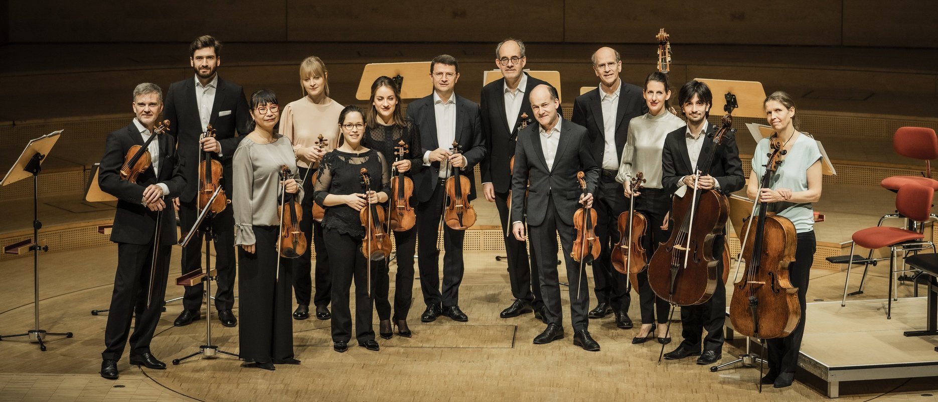 Zwölf Musiker in förmlicher Kleidung stehen auf einer hölzernen Bühne in einem Konzertsaal und halten Streichinstrumente, darunter Geigen, Bratschen und Celli, vor leeren Notenständern und Stühlen im Hintergrund.