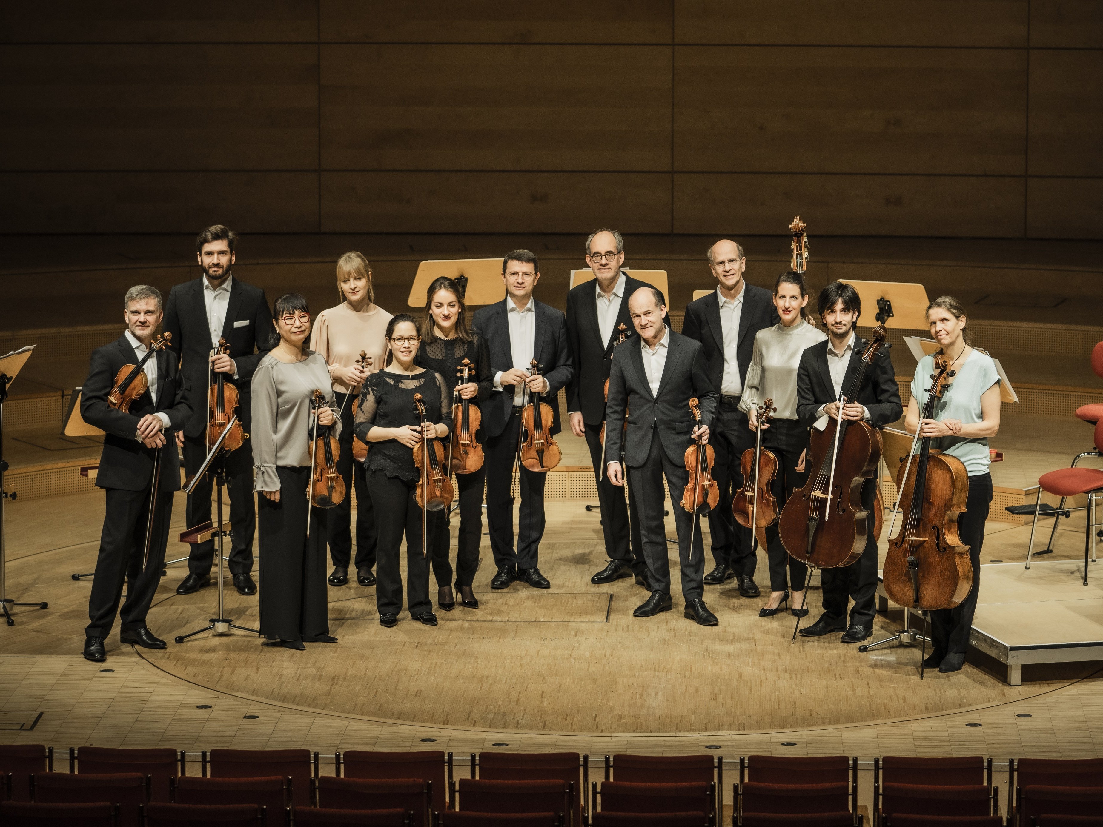 Zwölf Musiker in förmlicher Kleidung stehen auf einer hölzernen Bühne in einem Konzertsaal und halten Streichinstrumente, darunter Geigen, Bratschen und Celli, vor leeren Notenständern und Stühlen im Hintergrund.