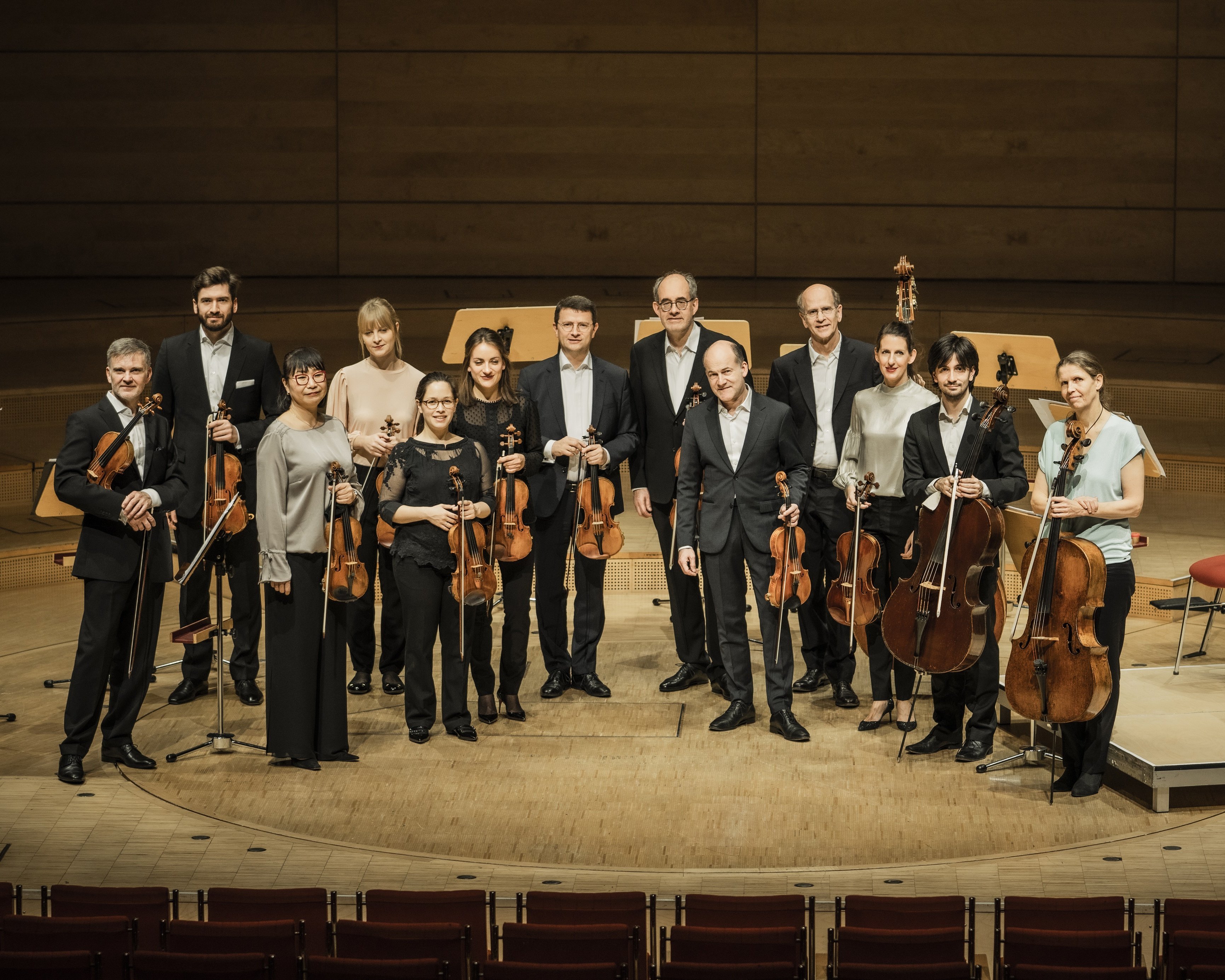 A group of thirteen musicians holding string instruments stands on a wooden concert stage, posing and smiling for a photo. Several empty chairs and music stands are visible in the background.