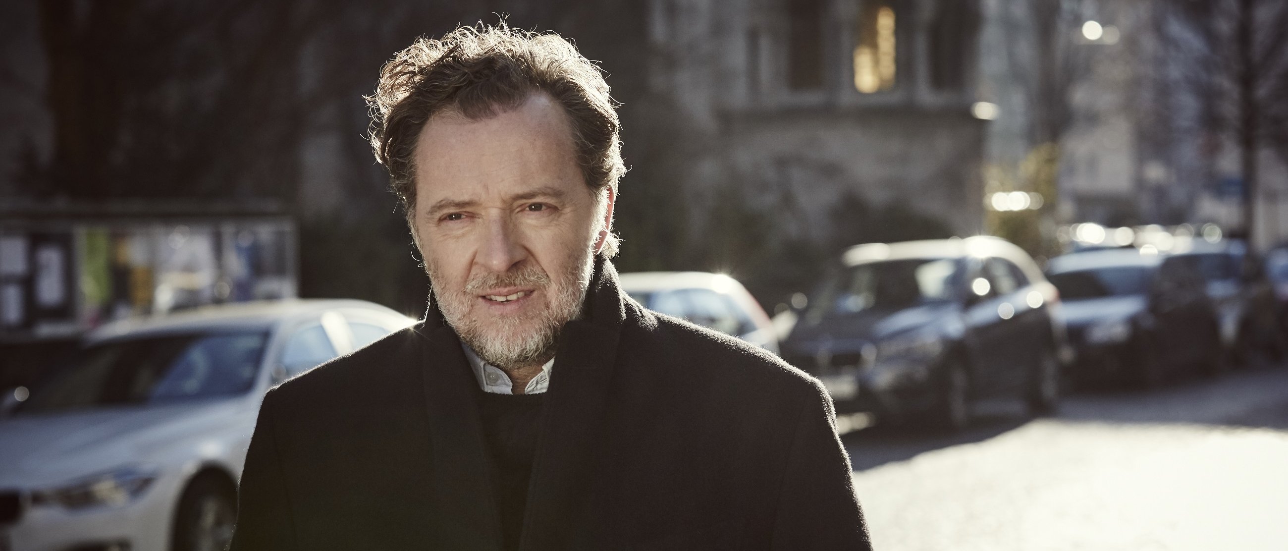 Christian Gerhaher mit gewelltem Haar und Bart, der einen dunklen Mantel trägt, steht im Freien auf einer sonnenbeschienenen Straße mit geparkten Autos und unscharfen Gebäuden im Hintergrund.